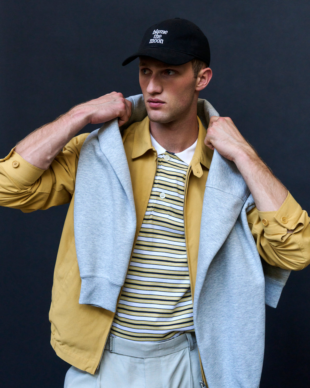 Man wearing the black Artless Blame the Moon organic cotton dad cap with a grey hood, striped polo and yellow jacket