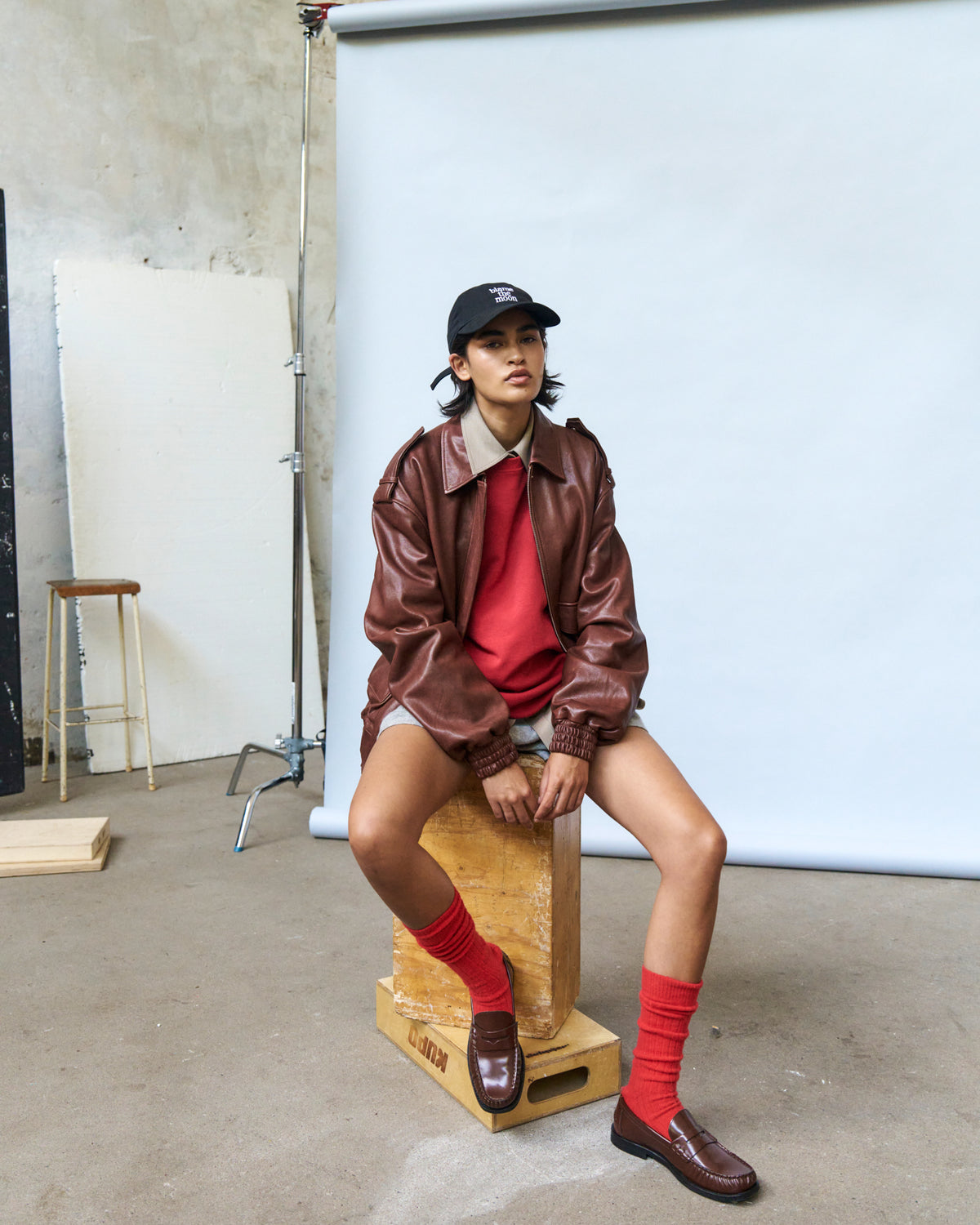 Person seated on a wooden block in a studio wearing the black Artless Blame the Moon organic cotton dad cap and a brown leather jacket