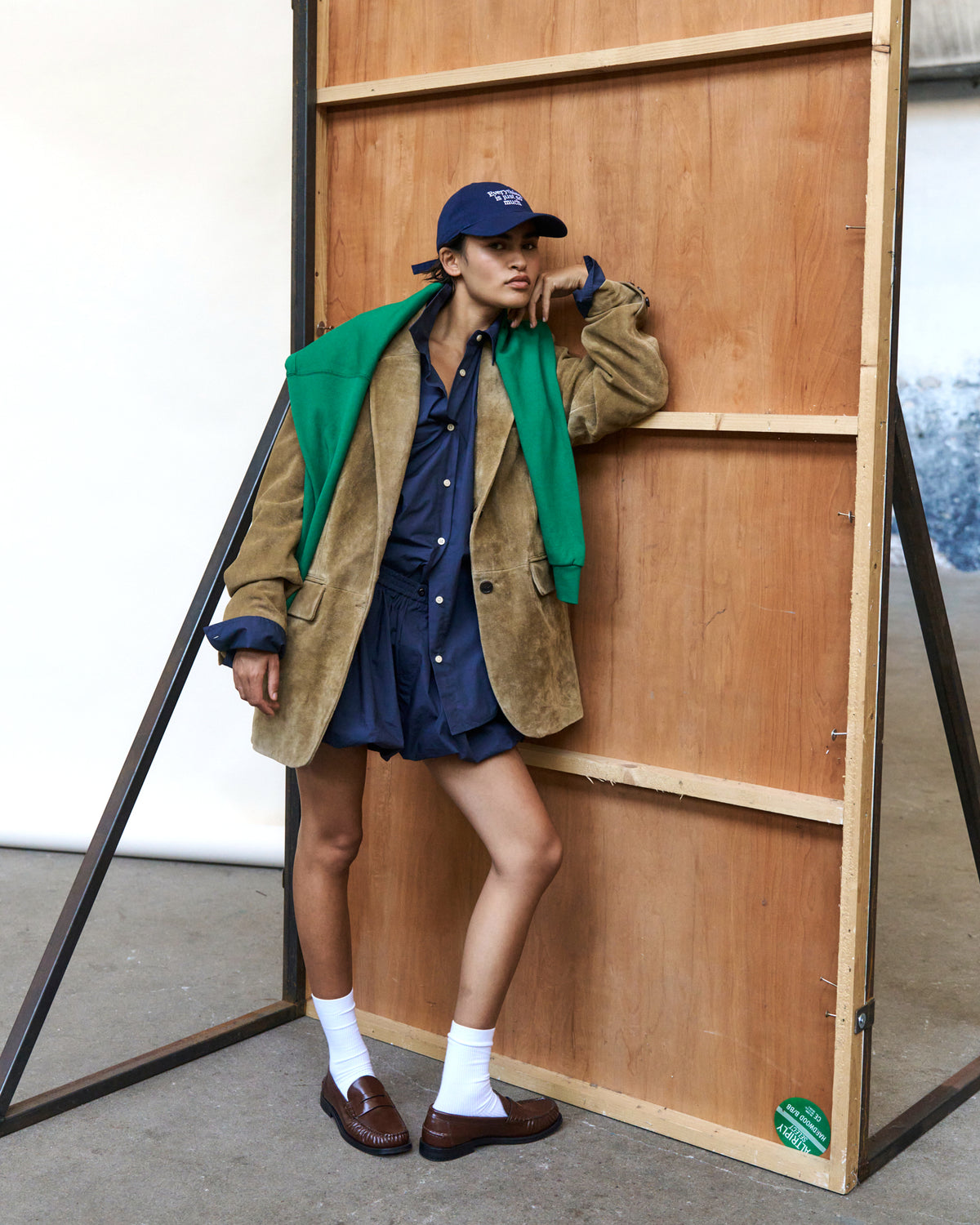 Person leaning on a wooden board wearing the navy Artless Everything Is Just So Much organic cotton dad cap, green jacket and shorts