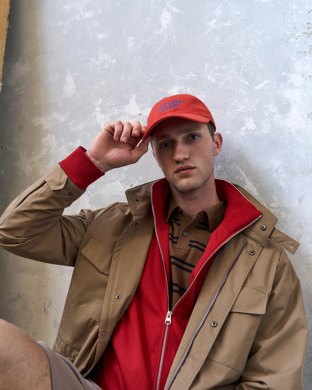 Man adjusting the red Artless Optimist Club organic cotton dad cap, tan jacket over a red hooded top