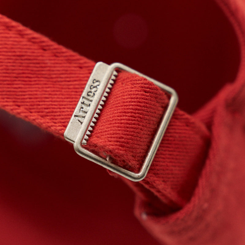 Close-up of the engraved Artless buckle on the red unstructured 6-panel dad cap made from GOTS certified organic cotton