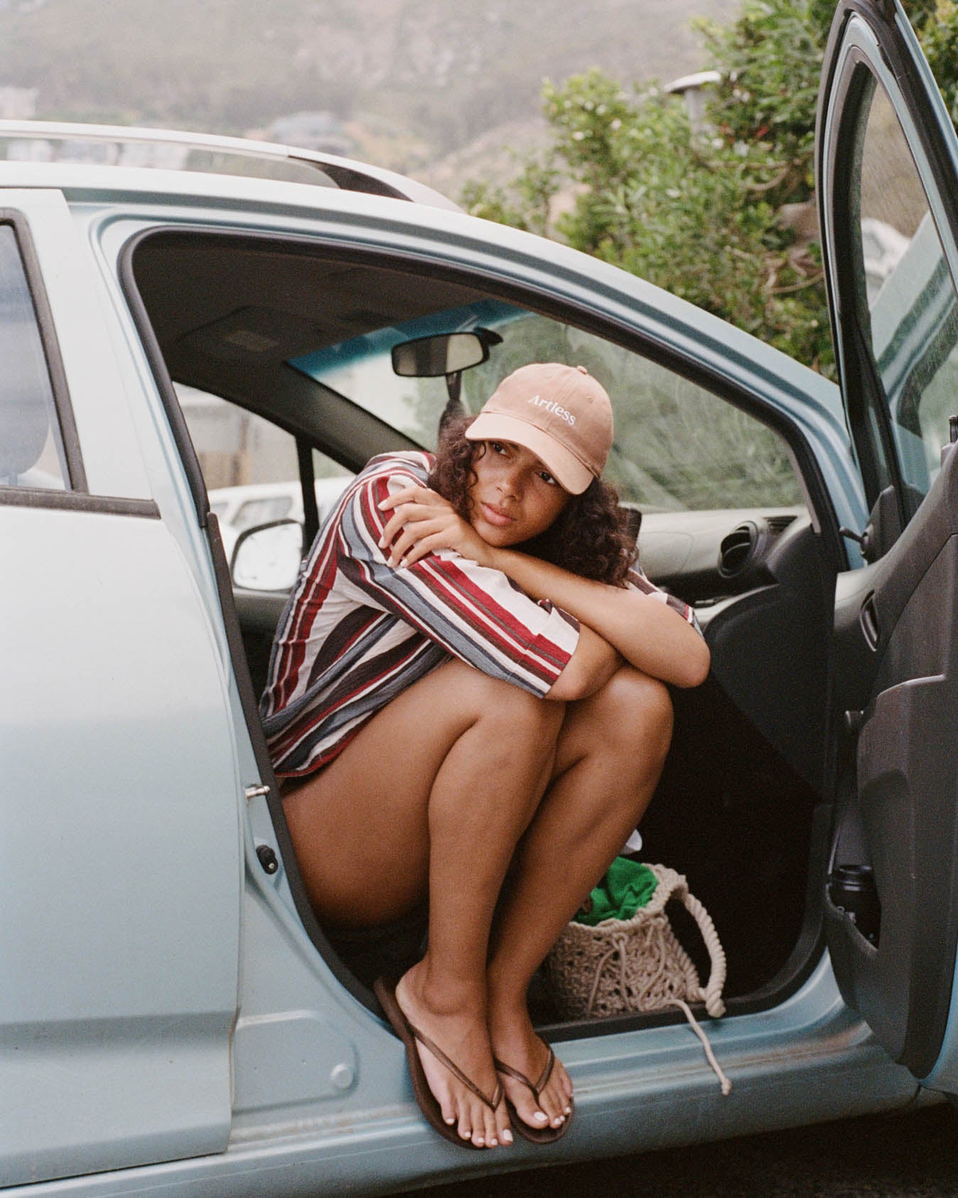 Woman sitting in the open door of a car wearing a brown unstructured 6-panel dad cap made from GOTS certified organic cotton by Artless