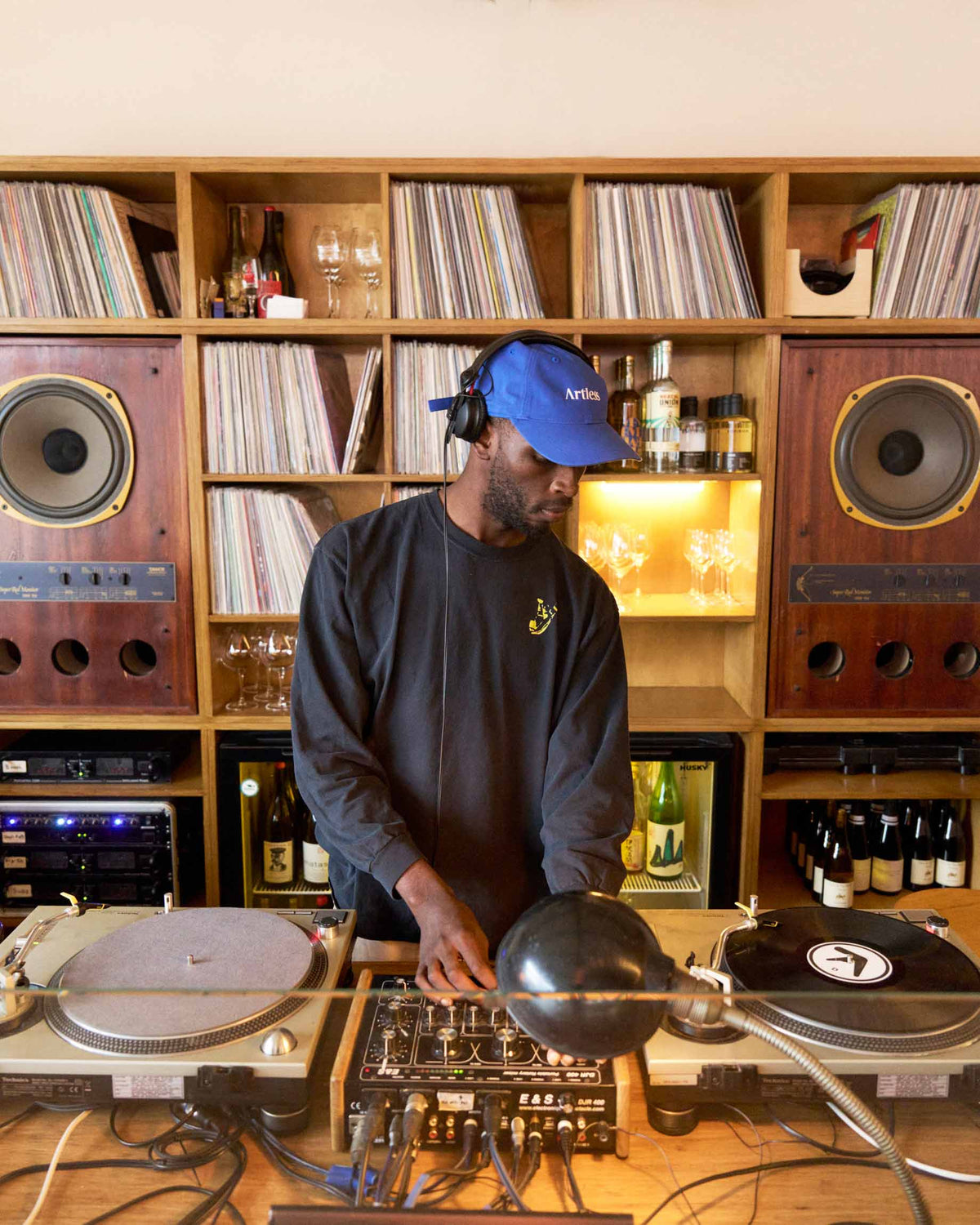 Person operating DJ equipment in a room with record players and shelves filled with records wearing a blue unstructured 6-panel dad cap made from GOTS certified organic cotton by Artless