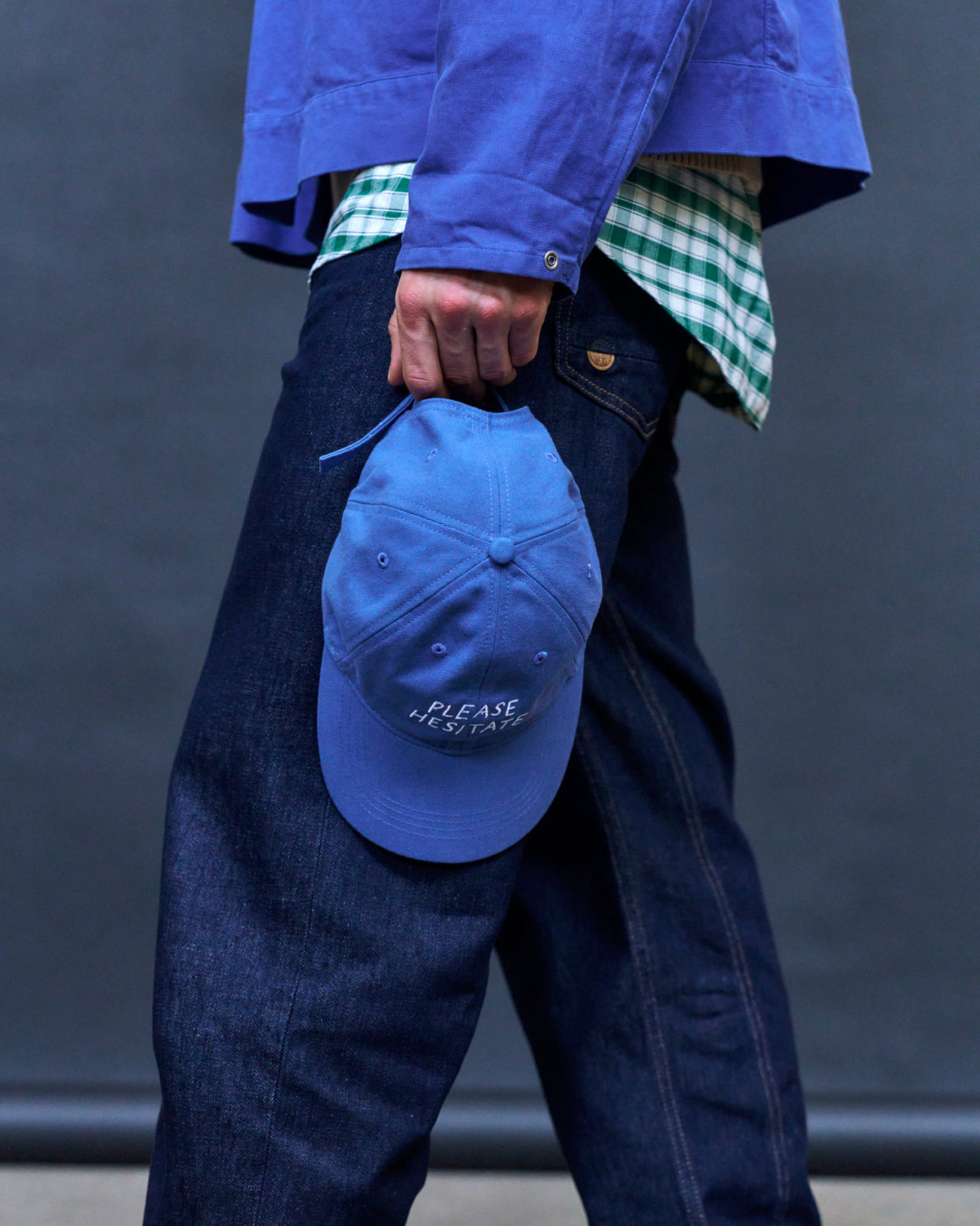 Person holding the blue Artless Please Hesitate organic cotton dad cap at their hip, blue jacket over a checked shirt and denim