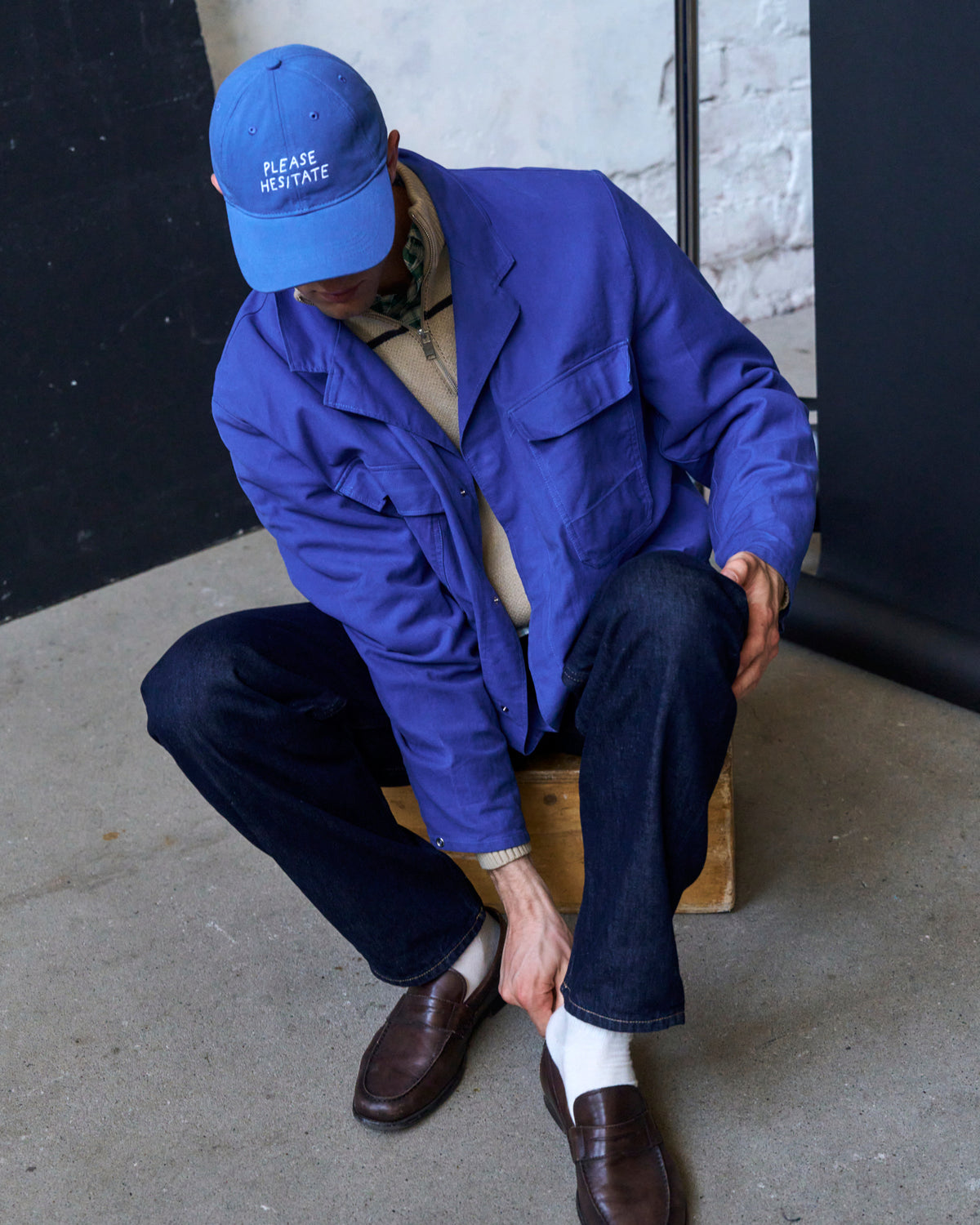 Man seated on a wooden box wearing the blue Artless Please Hesitate organic cotton dad cap, blue jacket and trousers, white socks