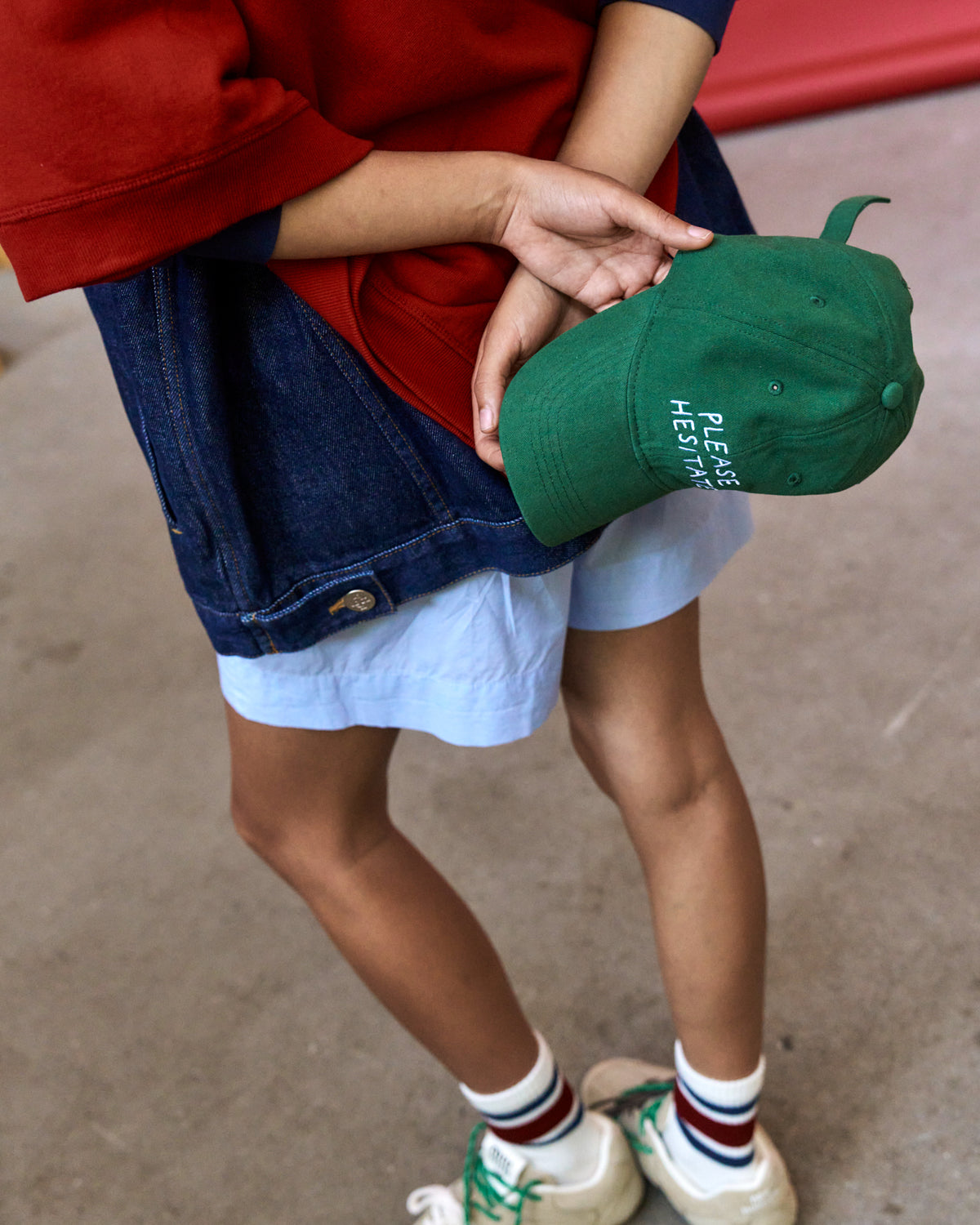 Person holding the green Artless Please Hesitate organic cotton dad cap at their hip, red top, denim jacket and wide jeans