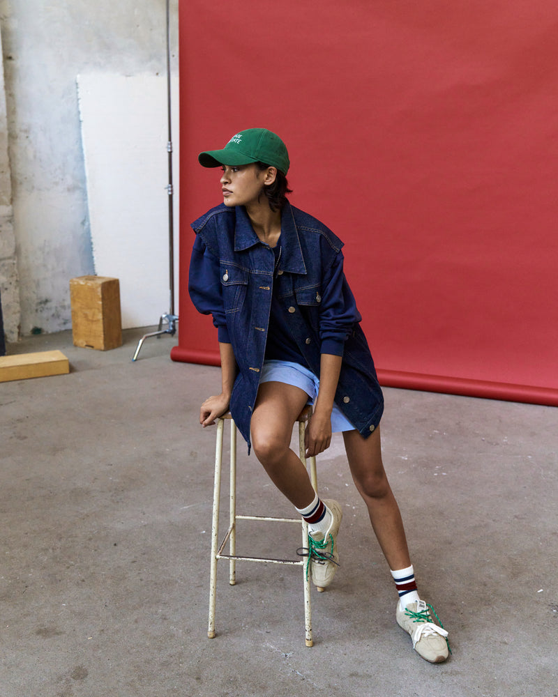 Person seated on a stool against a red wall wearing the green Artless Please Hesitate organic cotton dad cap and a denim jacket