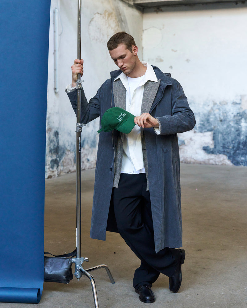 Man holding the green Artless Please Hesitate organic cotton dad cap, grey coat over a white shirt against a blue backdrop