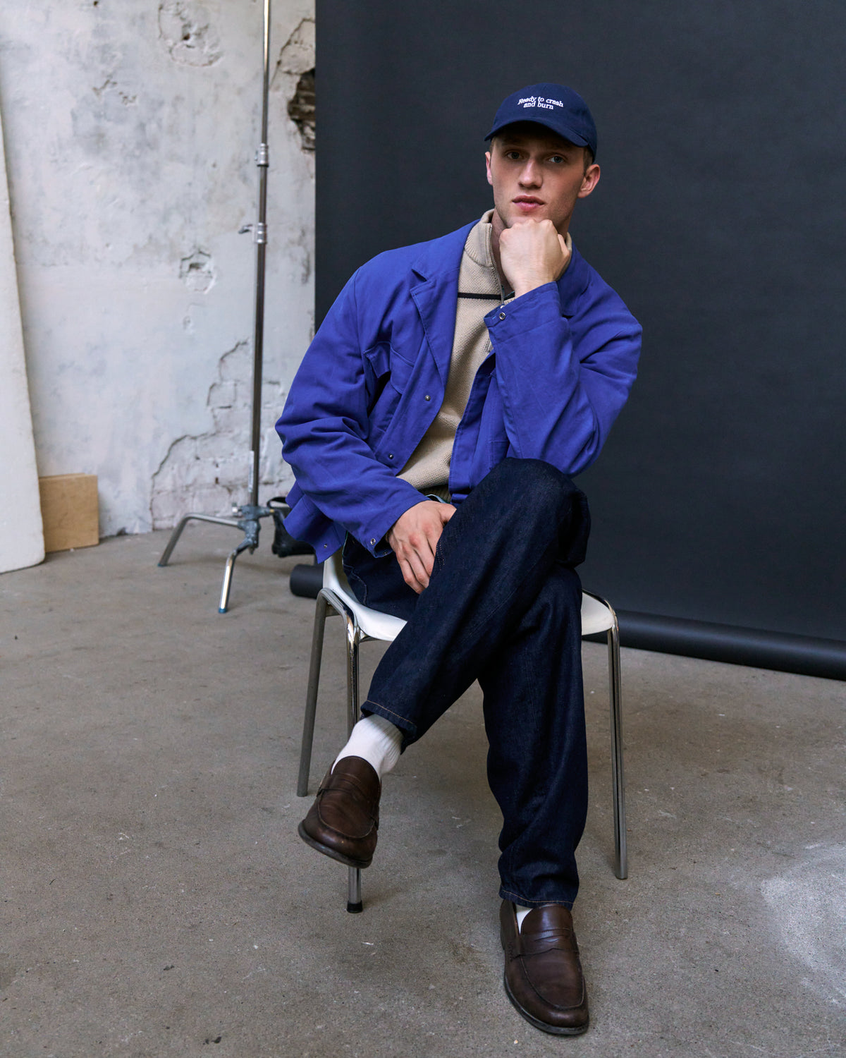 Man seated on a chair wearing the navy Artless Ready To Crash And Burn organic cotton dad cap, blue jacket over beige knit