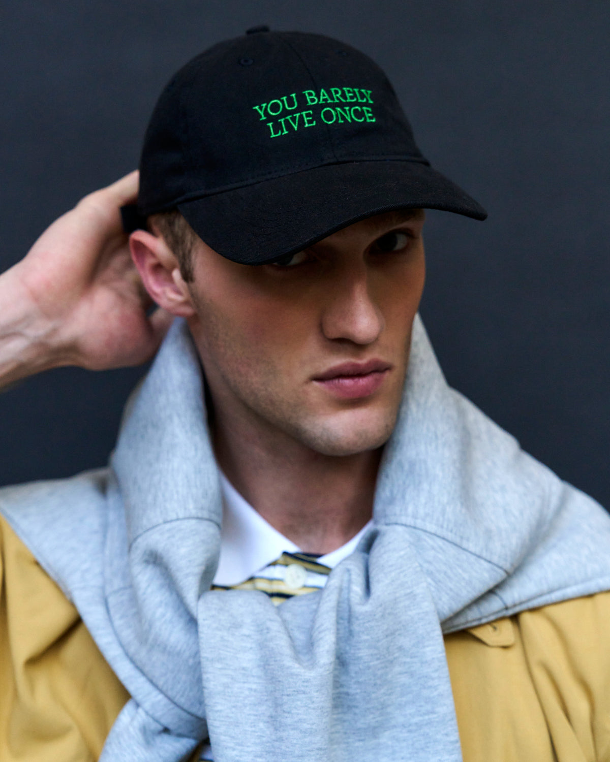 Portrait of a man wearing the black Artless You Barely Live Once organic cotton dad cap with green embroidery and a yellow jacket
