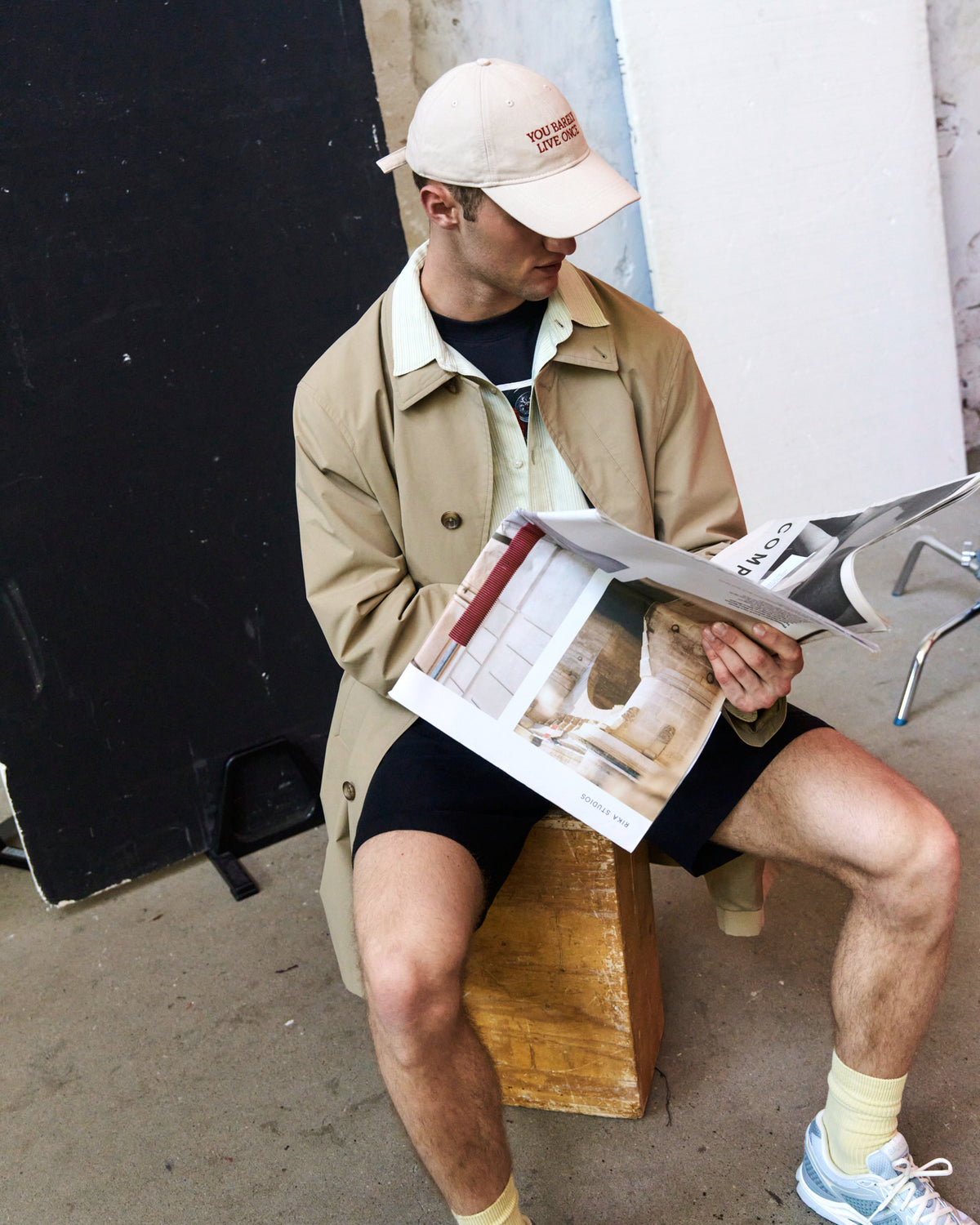 Man seated on a box reading a magazine while wearing the sand Artless You Barely Live Once organic cotton dad cap and a tan jacket