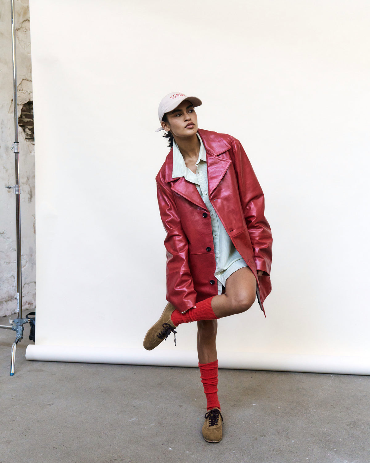 Woman against a white backdrop wearing the sand Artless You Barely Live Once organic cotton dad cap, red leather jacket and red socks