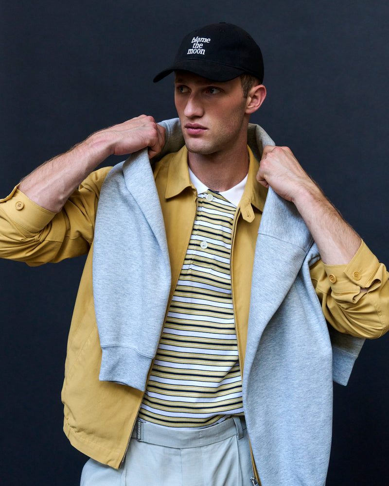 Man wearing the black Artless Blame the Moon organic cotton dad cap with a grey hood, striped polo and yellow jacket