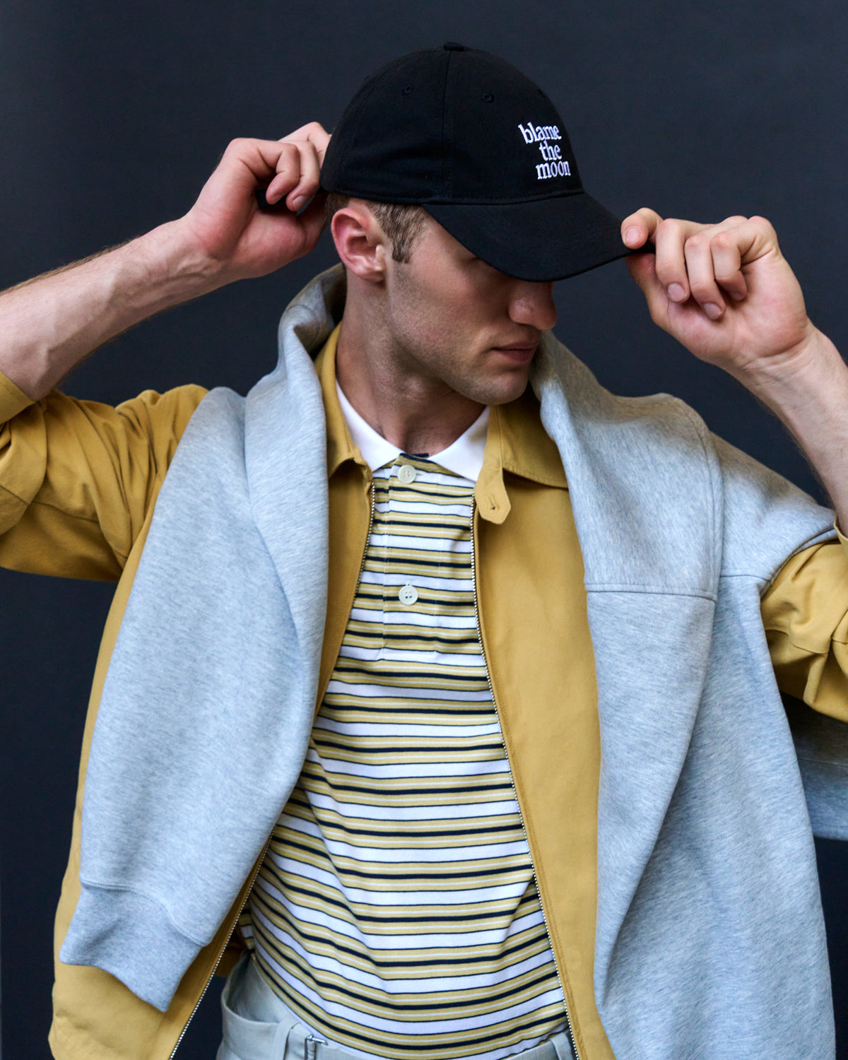 Man adjusting the black Artless Blame the Moon organic cotton dad cap, grey sweater layered over a yellow jacket
