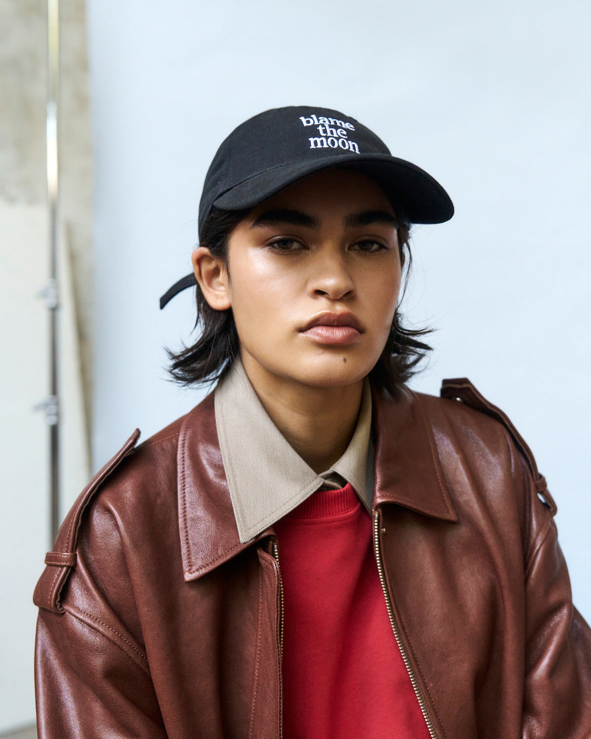 Portrait of a person wearing the black Artless Blame the Moon organic cotton dad cap with a brown leather jacket over a red top