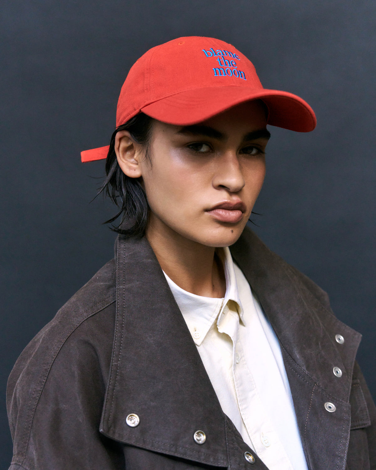 Portrait of a person wearing the red Artless Blame the Moon organic cotton dad cap with blue embroidery and a brown suede jacket