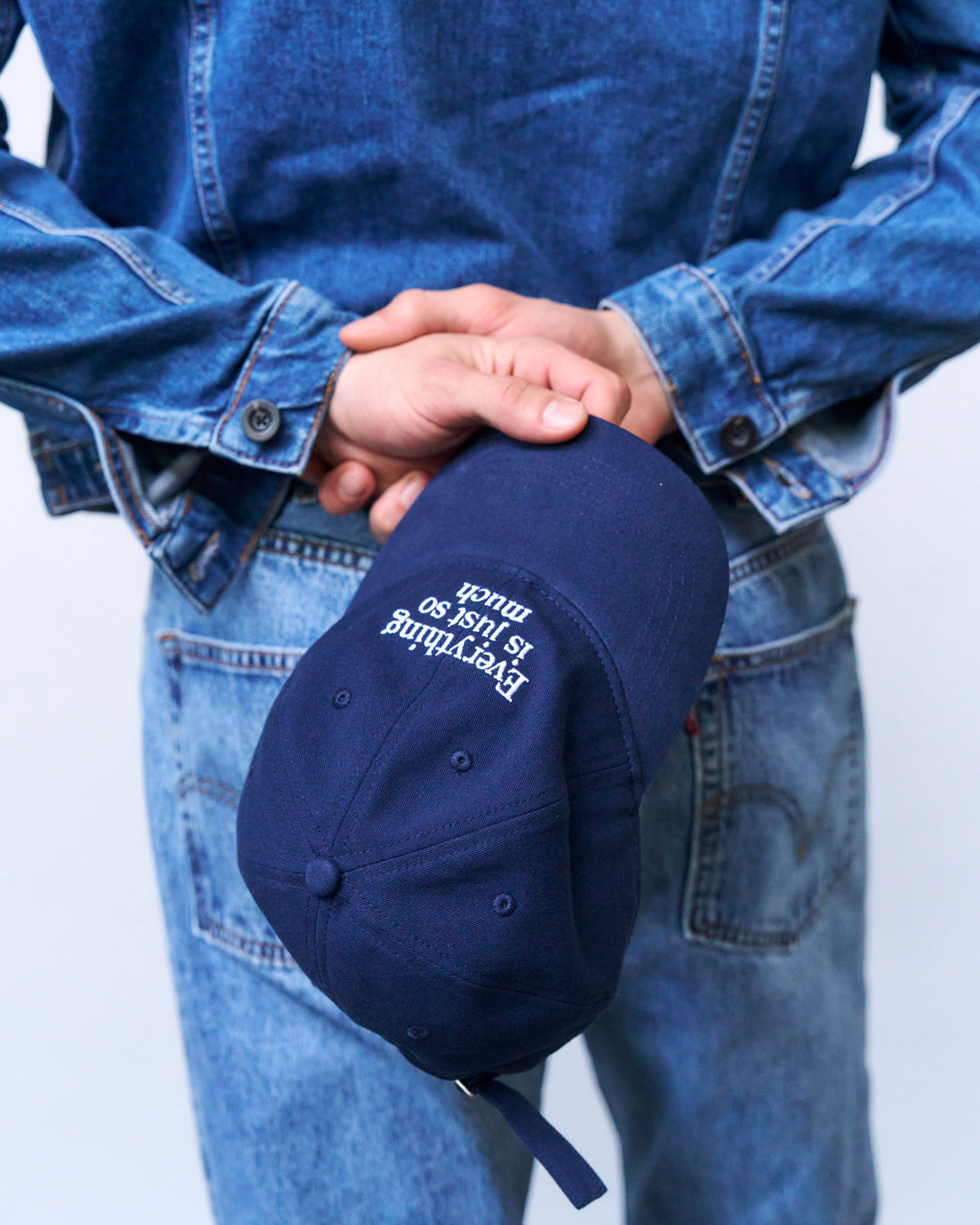 Close up of a person in double denim holding the navy Artless Everything Is Just So Much organic cotton dad cap at their hip