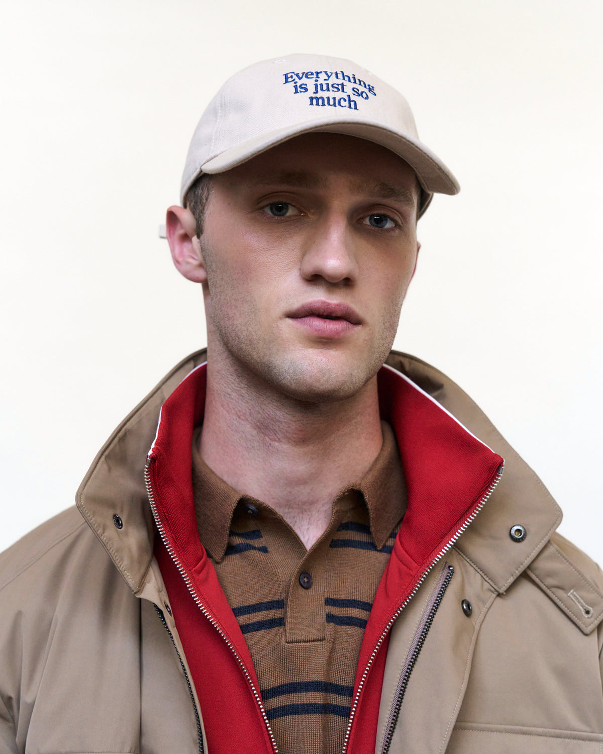 Portrait of a man wearing the sand Artless Everything Is Just So Much organic cotton dad cap with blue embroidery and a beige jacket