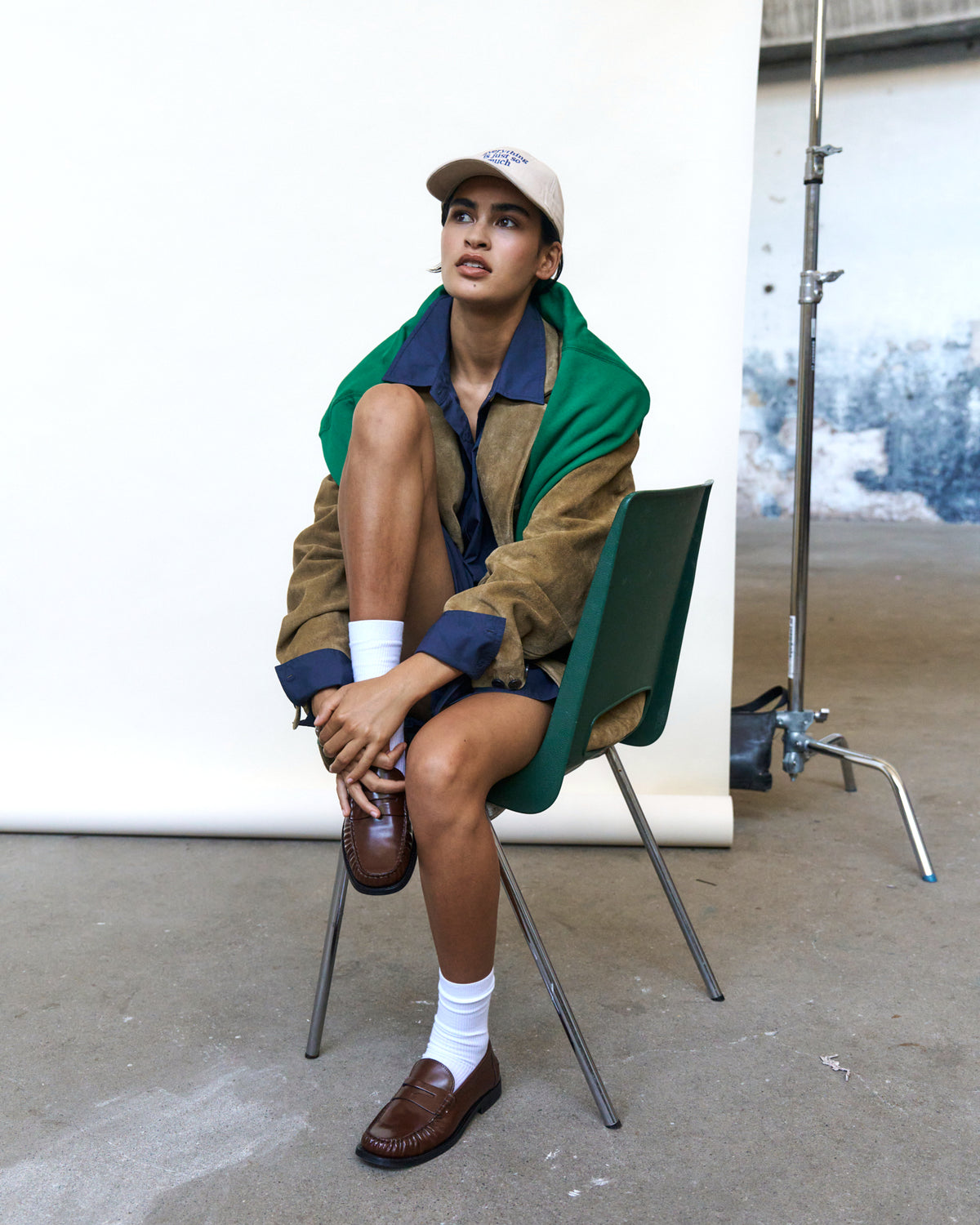 Person seated on a chair wearing the sand Artless Everything Is Just So Much organic cotton dad cap, green and brown jacket and loafers