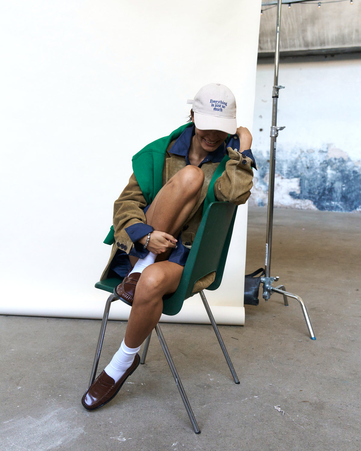 Person seated on a folding chair wearing the sand Artless Everything Is Just So Much organic cotton dad cap and a green and brown jacket