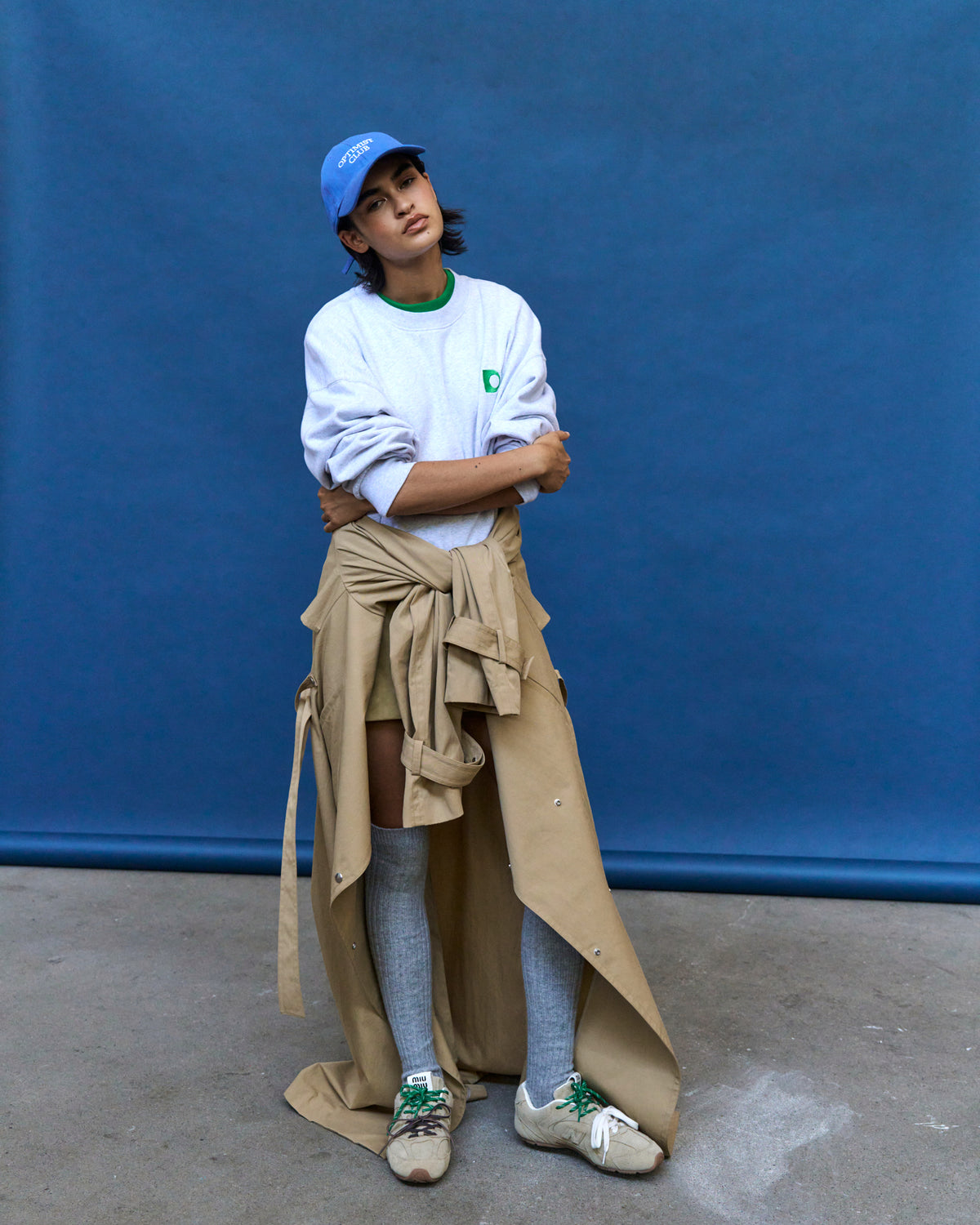 Person against a blue backdrop wearing the blue Artless Optimist Club organic cotton dad cap, white t-shirt and a trench tied at the waist