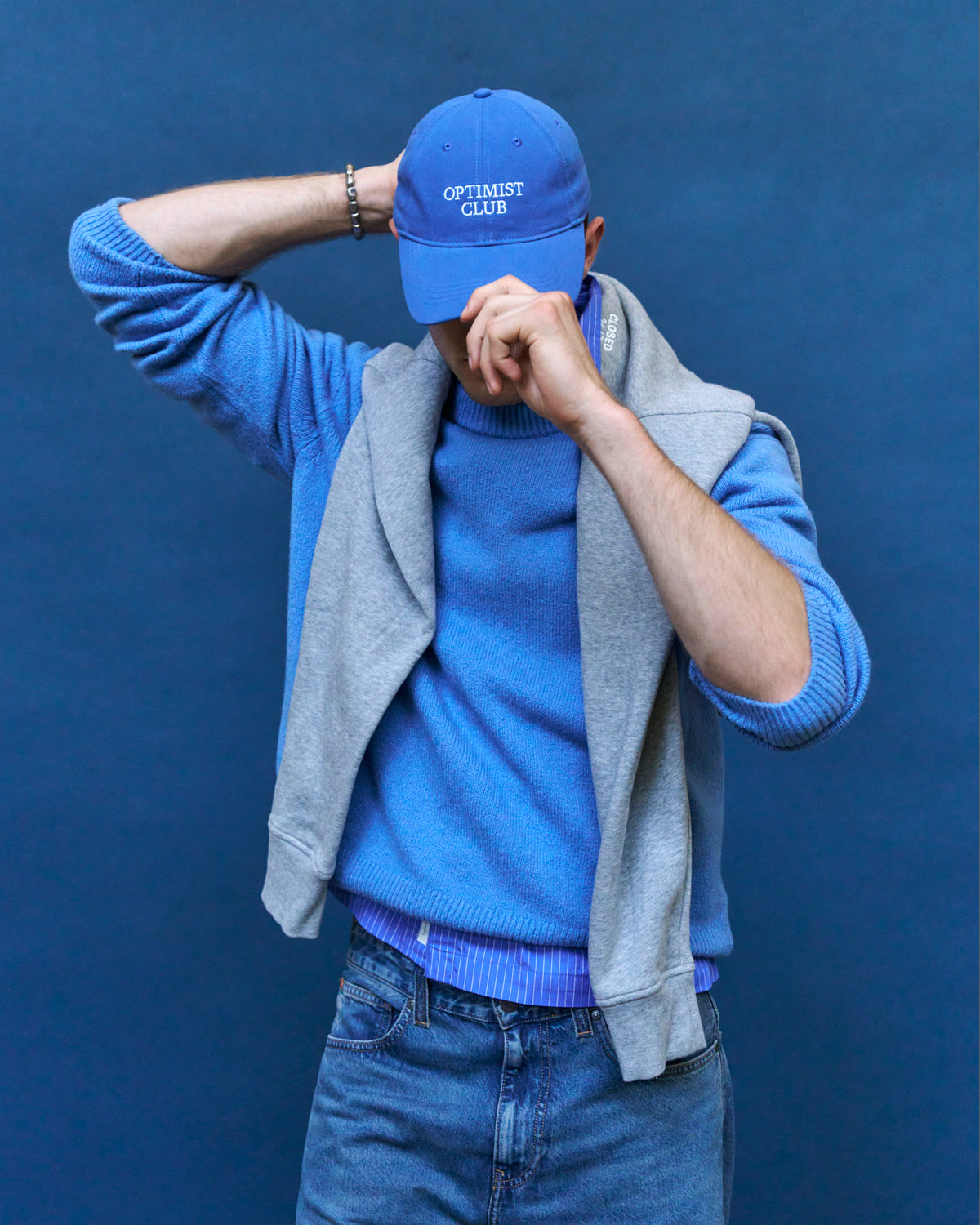 Person adjusting the blue Artless Optimist Club organic cotton dad cap, grey hoodie over a blue sweater and denim