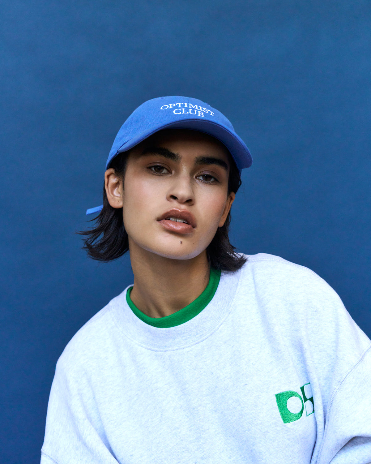 Portrait of a woman wearing the blue Artless Optimist Club organic cotton dad cap and a grey sweatshirt with green trim