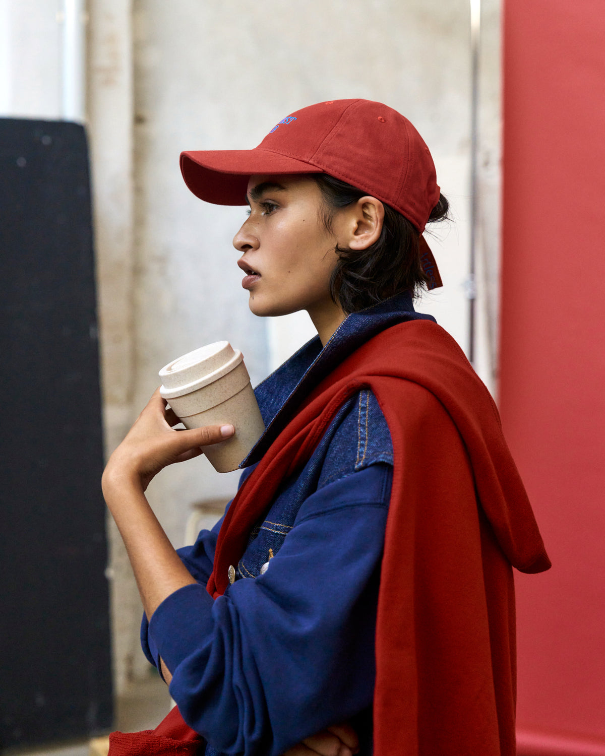 Person in profile holding a coffee cup while wearing the red Artless Optimist Club organic cotton dad cap and a red coat over navy