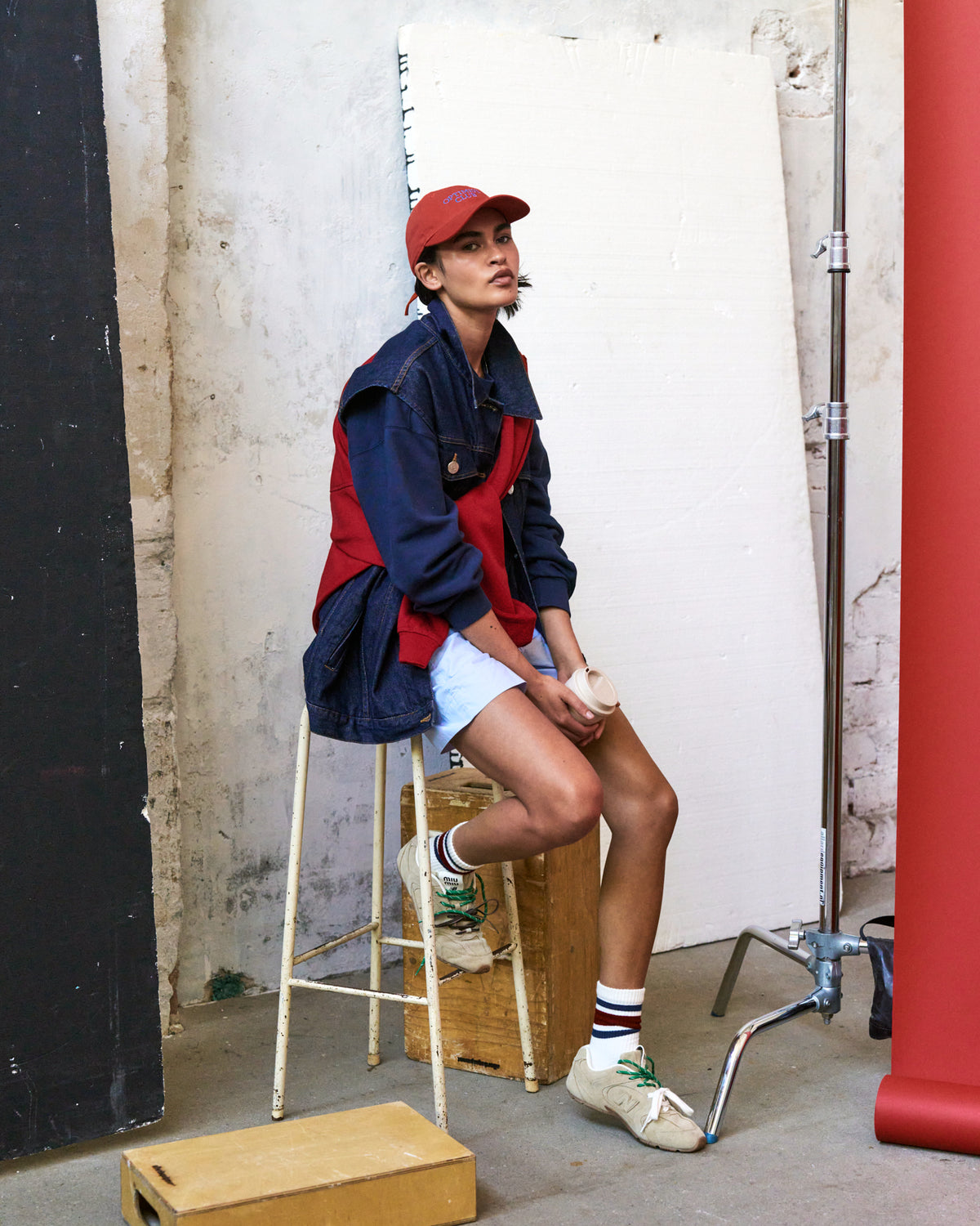 Person seated on a stool wearing the red Artless Optimist Club organic cotton dad cap, navy and red jacket and striped socks