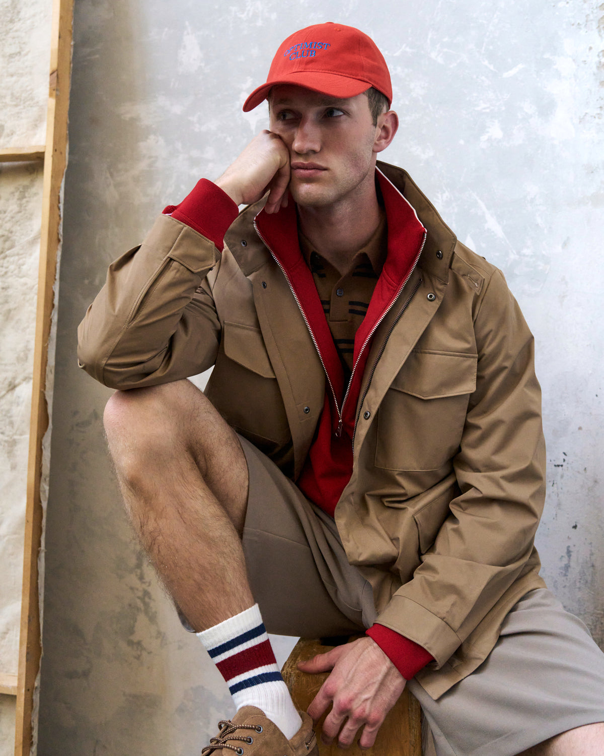 Man seated wearing the red Artless Optimist Club organic cotton dad cap, tan jacket and striped socks