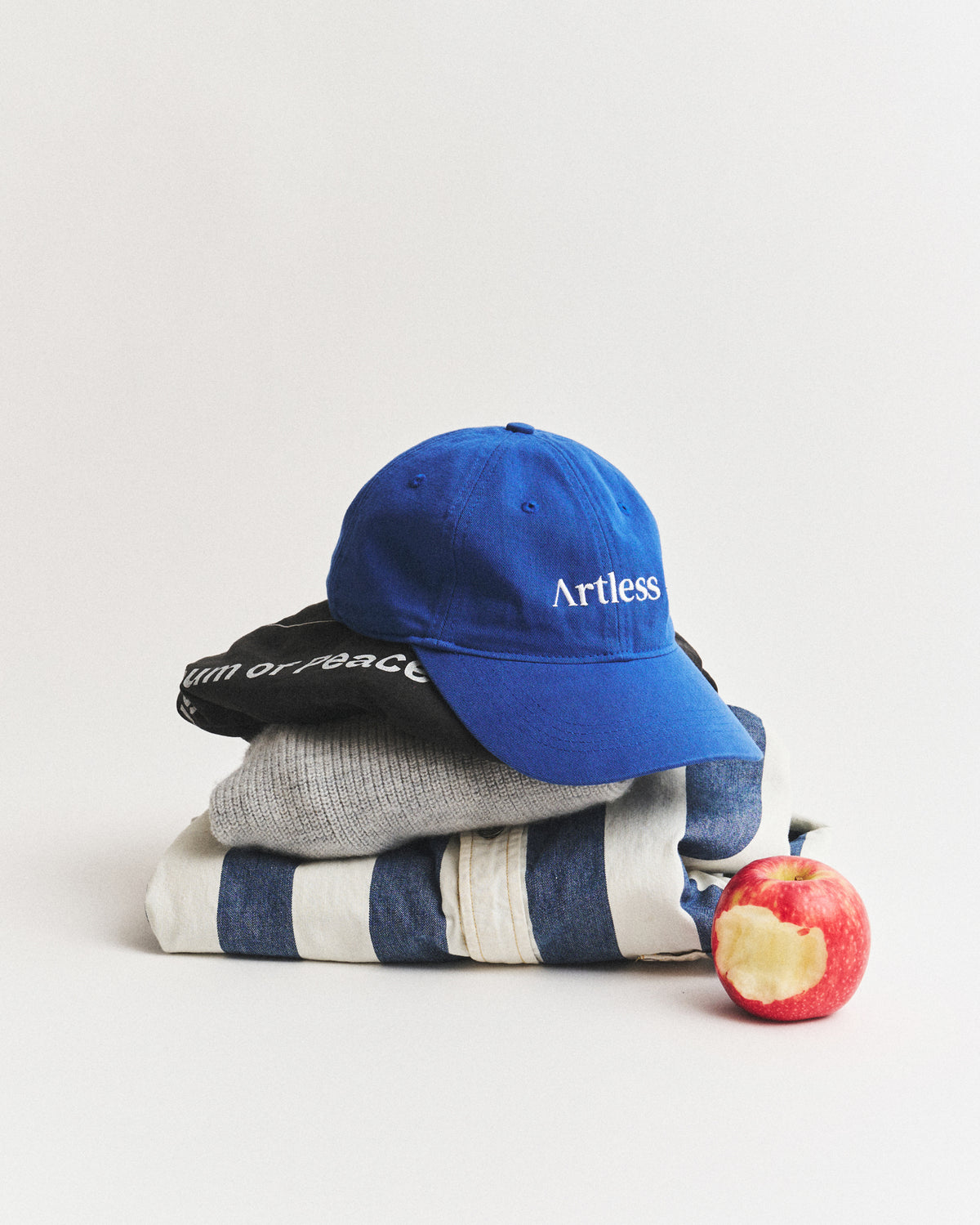 Blue Artless logo dad cap in GOTS certified organic cotton resting on a folded stack of knitwear beside a red apple