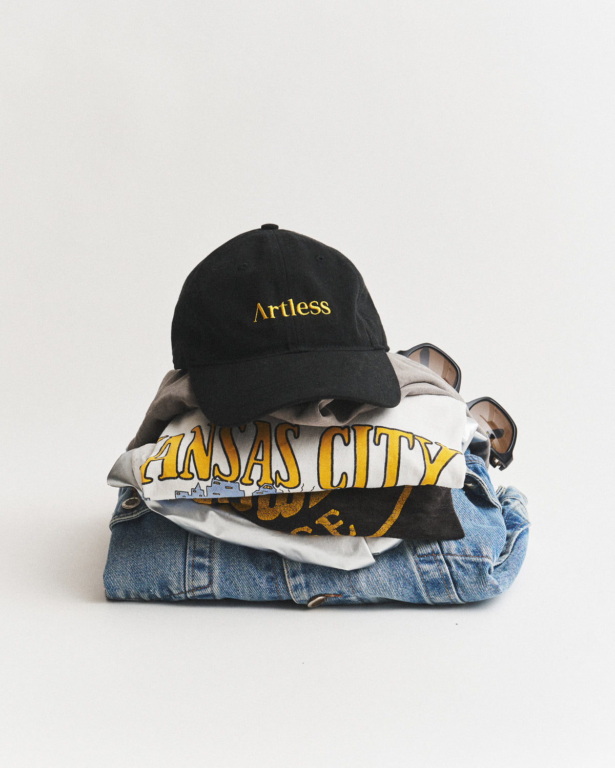 Black Artless logo dad cap in GOTS certified organic cotton resting on a folded stack of t-shirts and denim with sunglasses