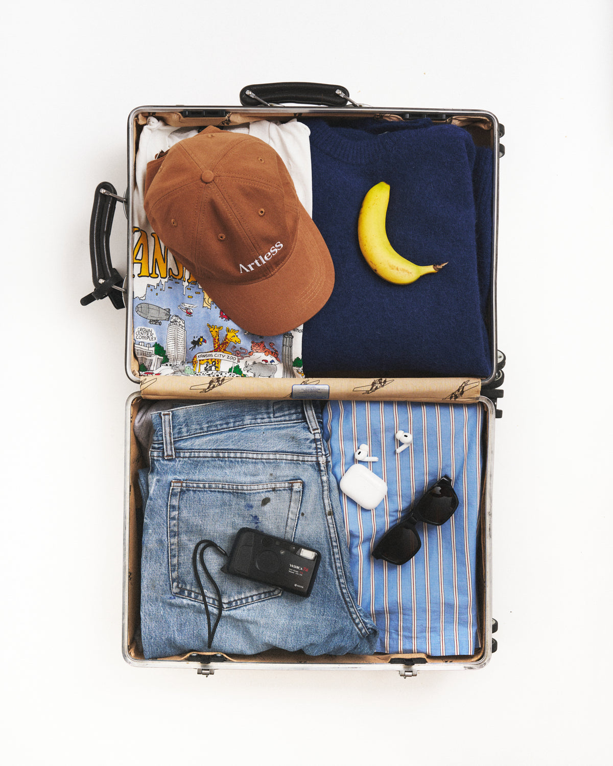 Open suitcase packed with travel essentials and the brown Artless organic cotton logo dad cap, flat lay on a light background