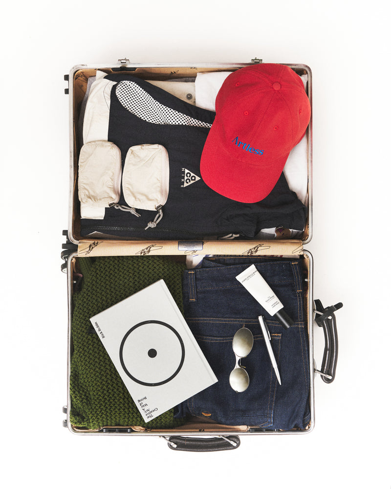 Open suitcase packed with travel essentials, a vinyl record and the red Artless organic cotton logo dad cap, flat lay