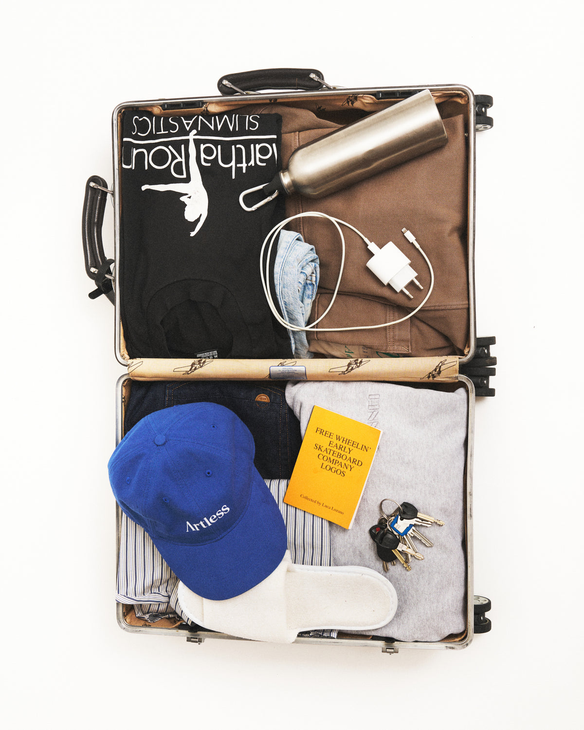 Open suitcase packed with travel essentials and the blue Artless organic cotton logo dad cap, flat lay on a light background