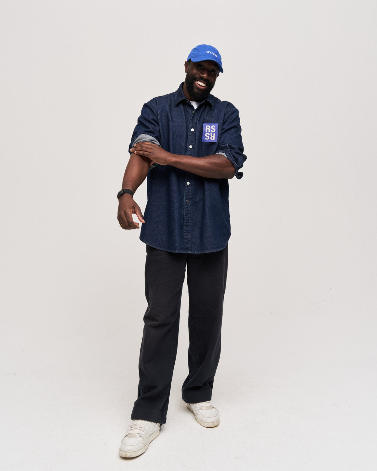 Man standing with arms crossed wearing the blue Artless organic cotton logo dad cap, blue shirt and dark trousers