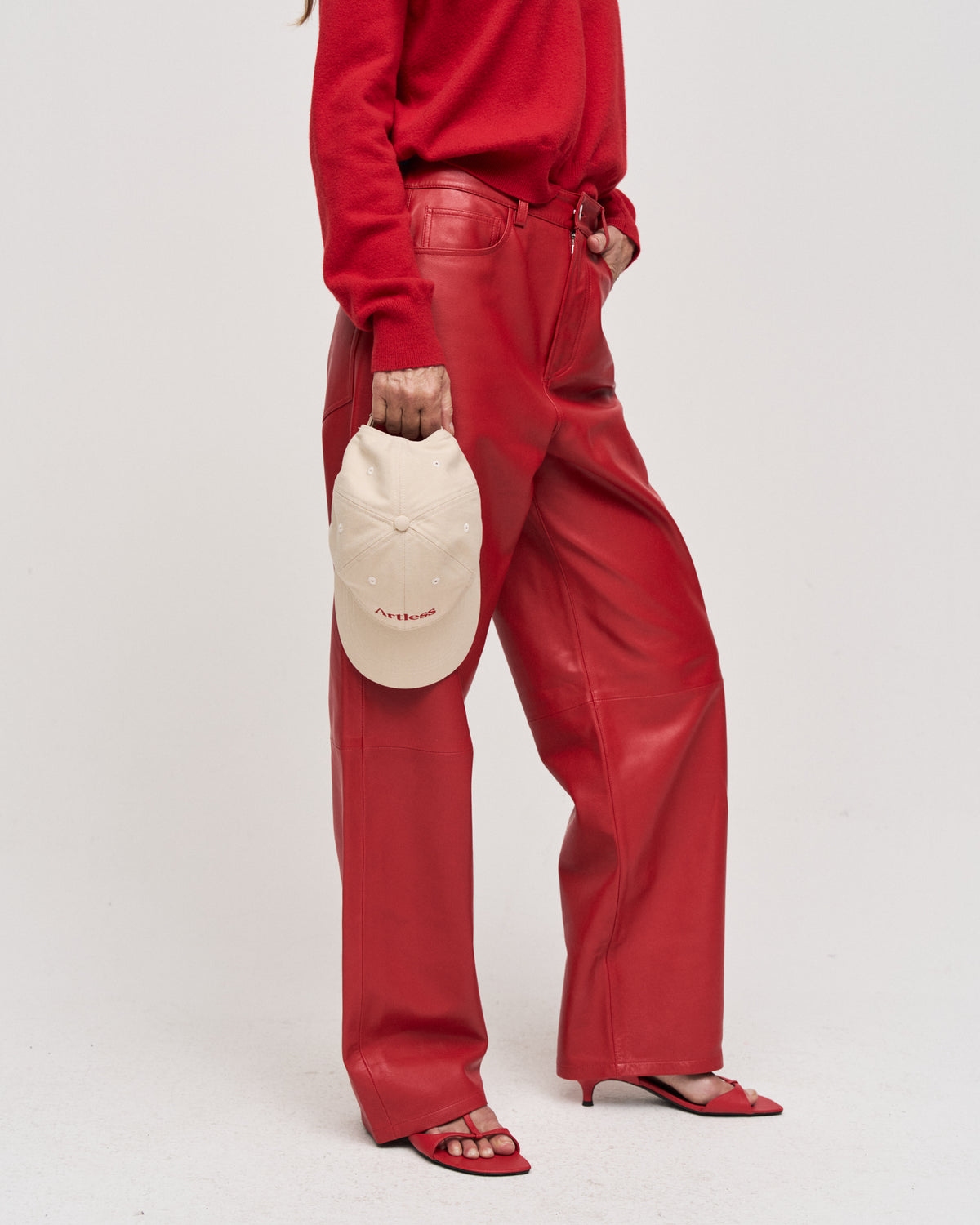 Person in red leather trousers and heels holding the sand Artless organic cotton logo dad cap at their side