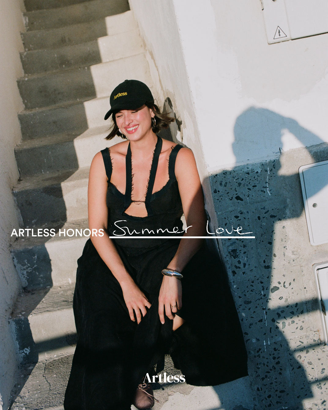 Woman in black dress and cap sitting on steps wearing a black unstructured 6-panel dad cap made from GOTS certified organic cotton by Artless
