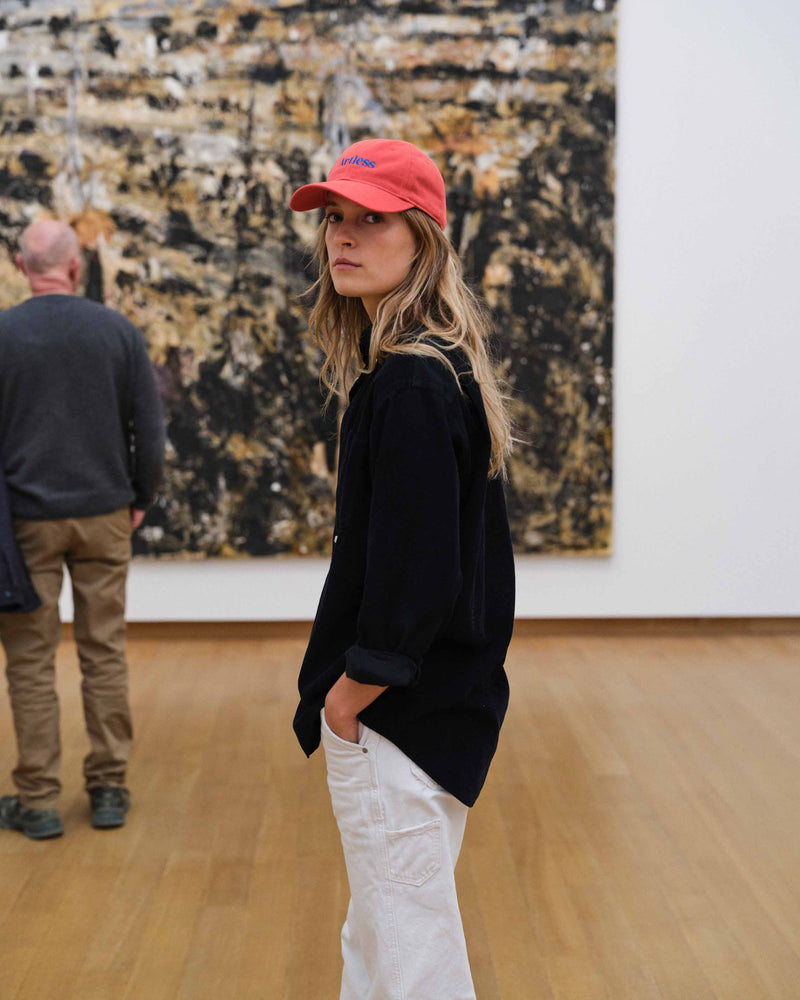 Woman wearing a red unstructured 6-panel dad cap made from GOTS certified organic cotton by Artless standing in an art gallery with abstract paintings on the wall.