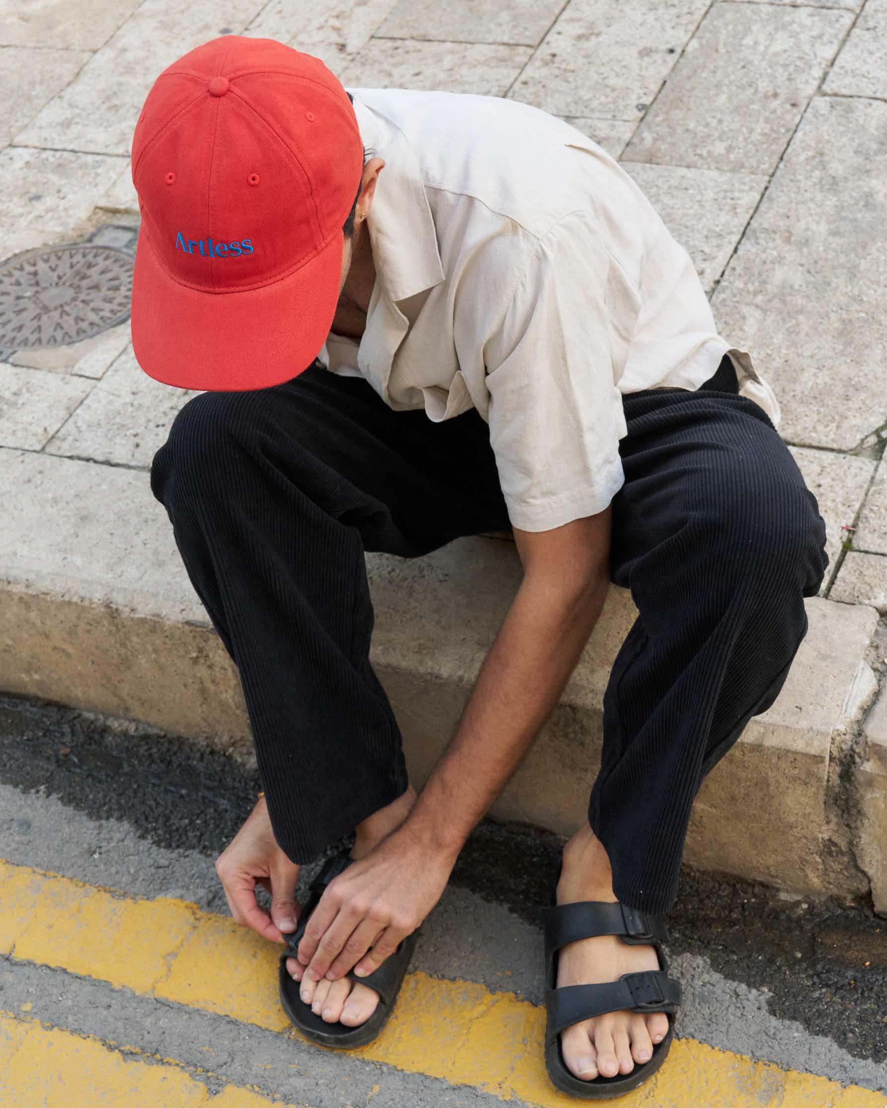 Person wearing a red unstructured 6-panel dad cap made from GOTS certified organic cotton by Artless sitting on a sidewalk