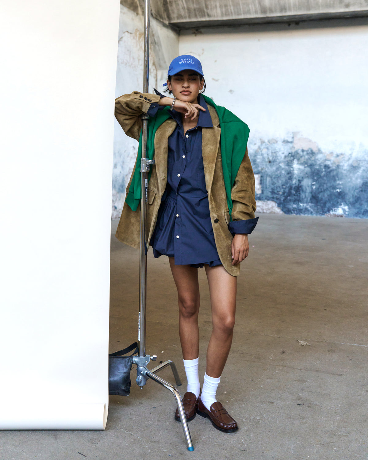 Person against a white backdrop wearing the blue Artless Please Hesitate organic cotton dad cap, green and tan jacket over a navy shirtdress