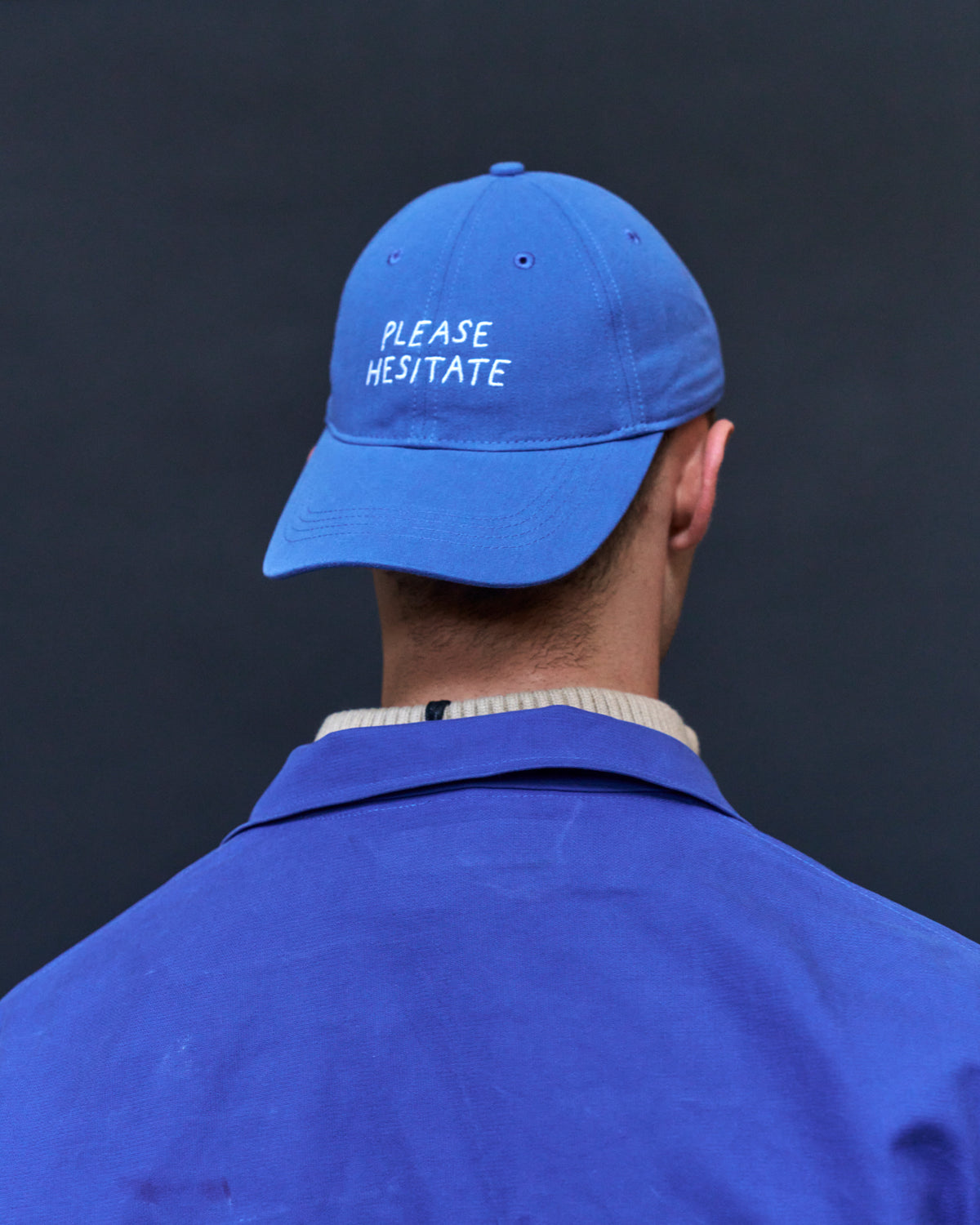 Back view of a person wearing the blue Artless Please Hesitate organic cotton dad cap with white embroidery and a blue jacket