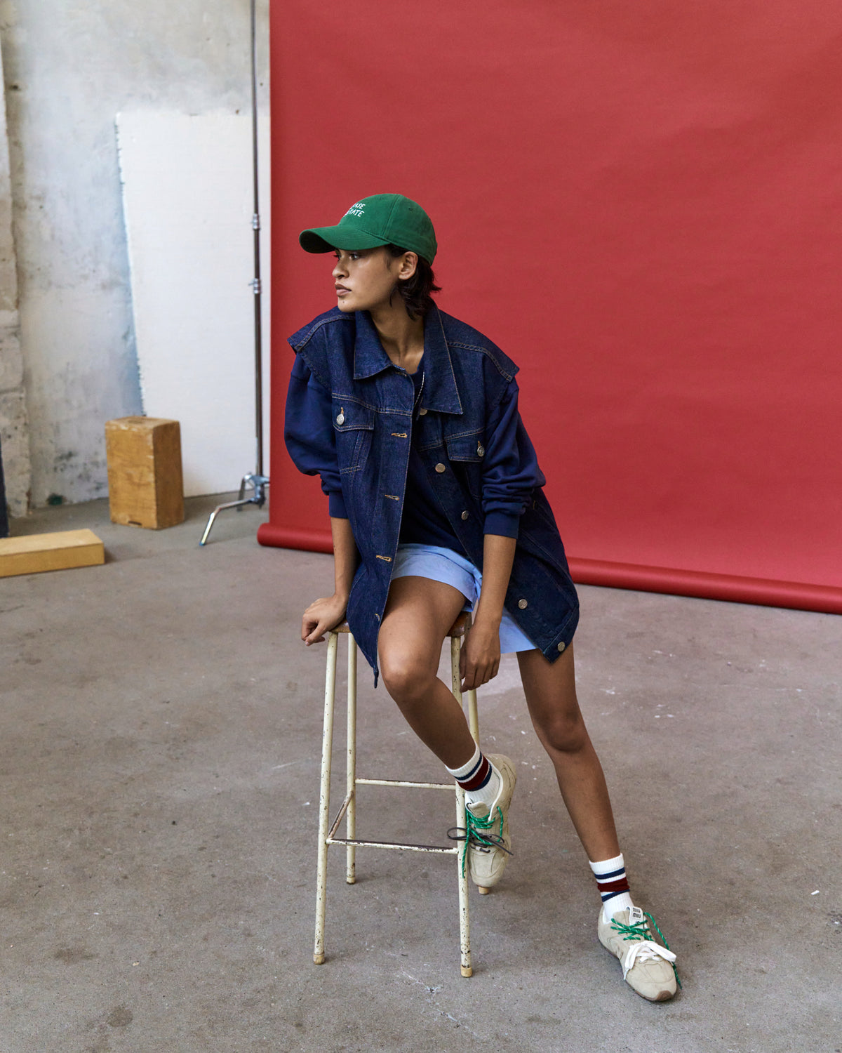 Person seated on a stool against a red wall wearing the green Artless Please Hesitate organic cotton dad cap and a denim jacket