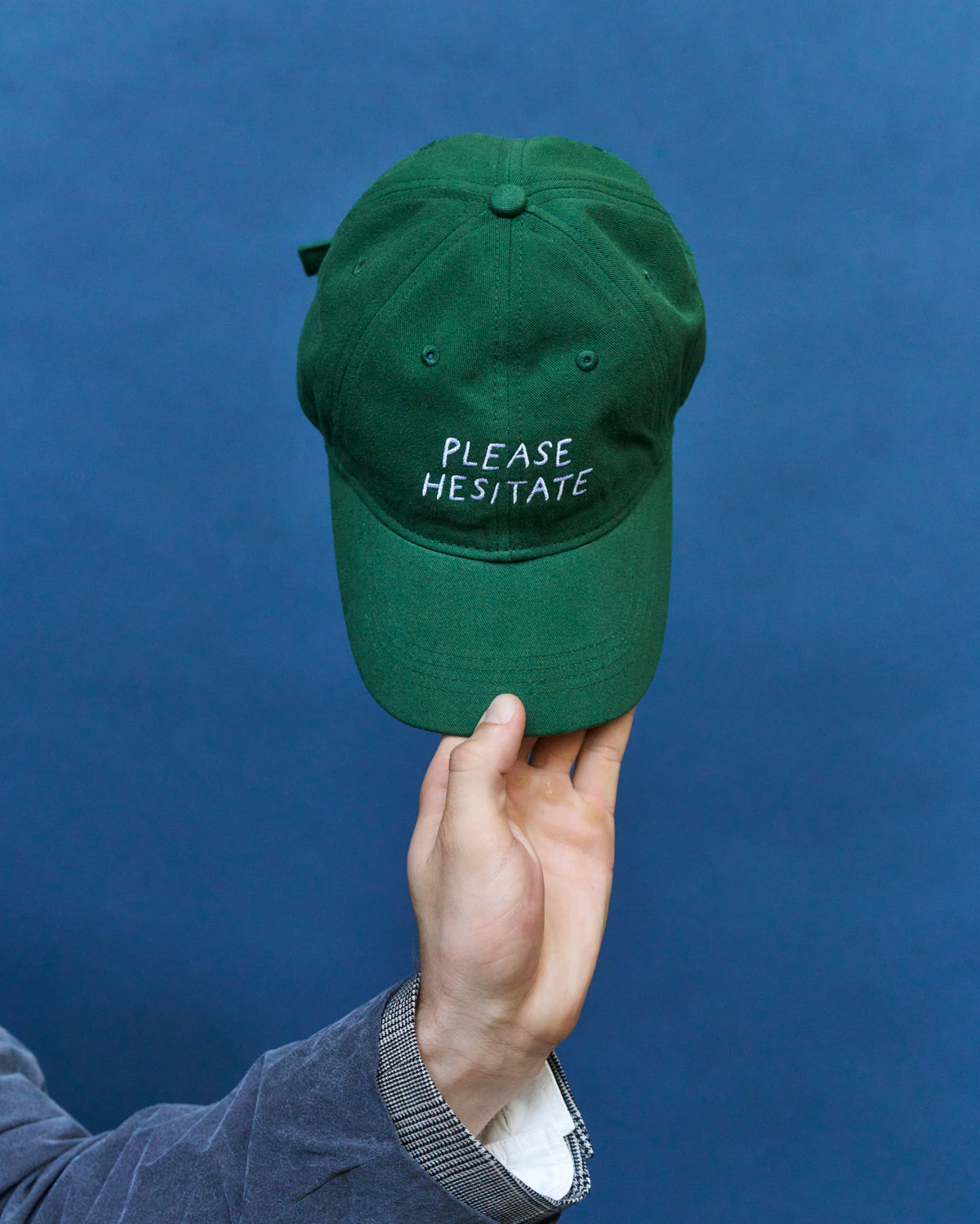 Hand holding up the green Artless Please Hesitate organic cotton dad cap with white embroidery against a blue backdrop