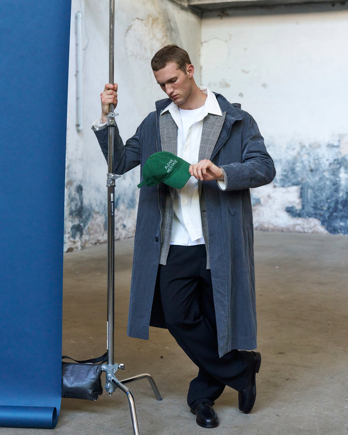 Man holding the green Artless Please Hesitate organic cotton dad cap, grey coat over a white shirt against a blue backdrop