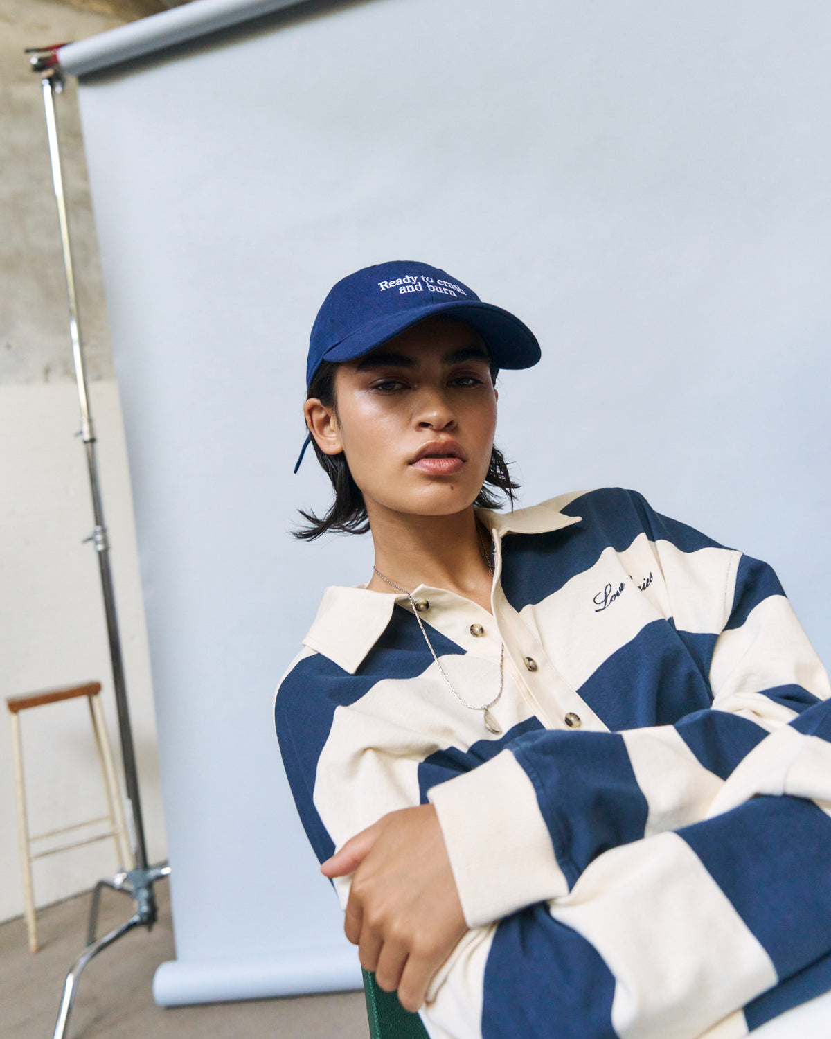 Woman leaning on a white backdrop roll wearing the navy Artless Ready To Crash And Burn organic cotton dad cap and a striped rugby shirt