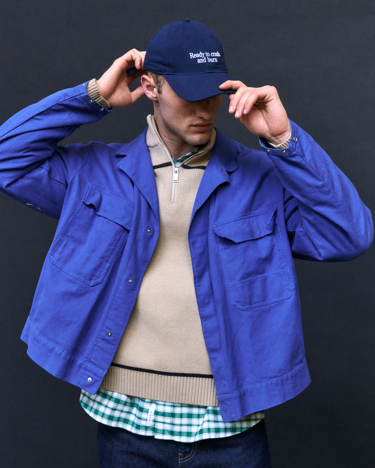 Man adjusting the navy Artless Ready To Crash And Burn organic cotton dad cap, blue jacket over a beige knit and checked shirt