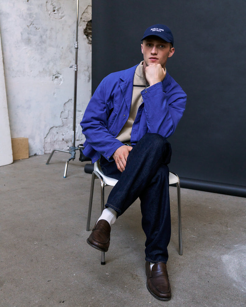 Man seated on a chair wearing the navy Artless Ready To Crash And Burn organic cotton dad cap, blue jacket over beige knit