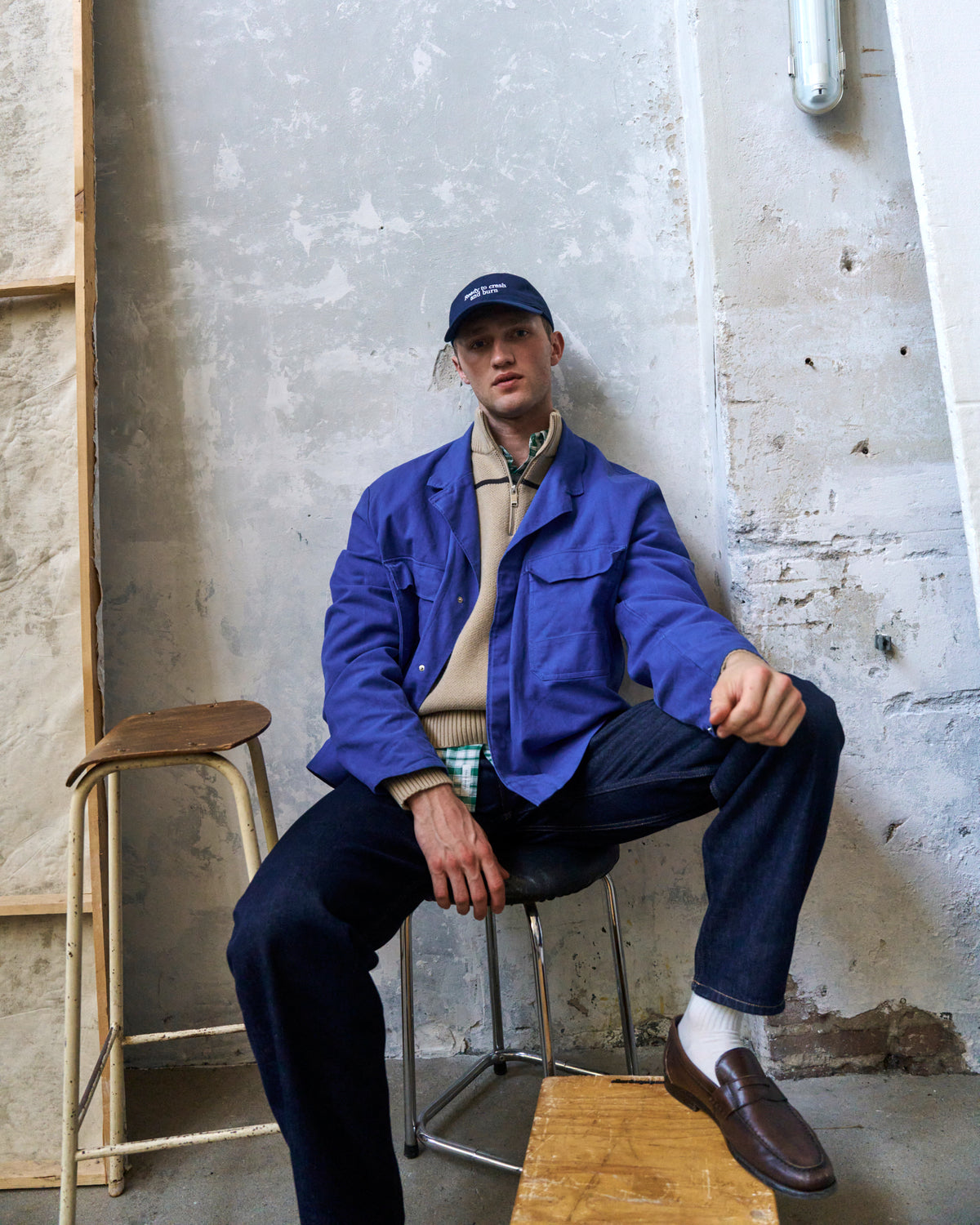 Man seated on a stool against a concrete wall wearing the navy Artless Ready To Crash And Burn organic cotton dad cap and a blue jacket