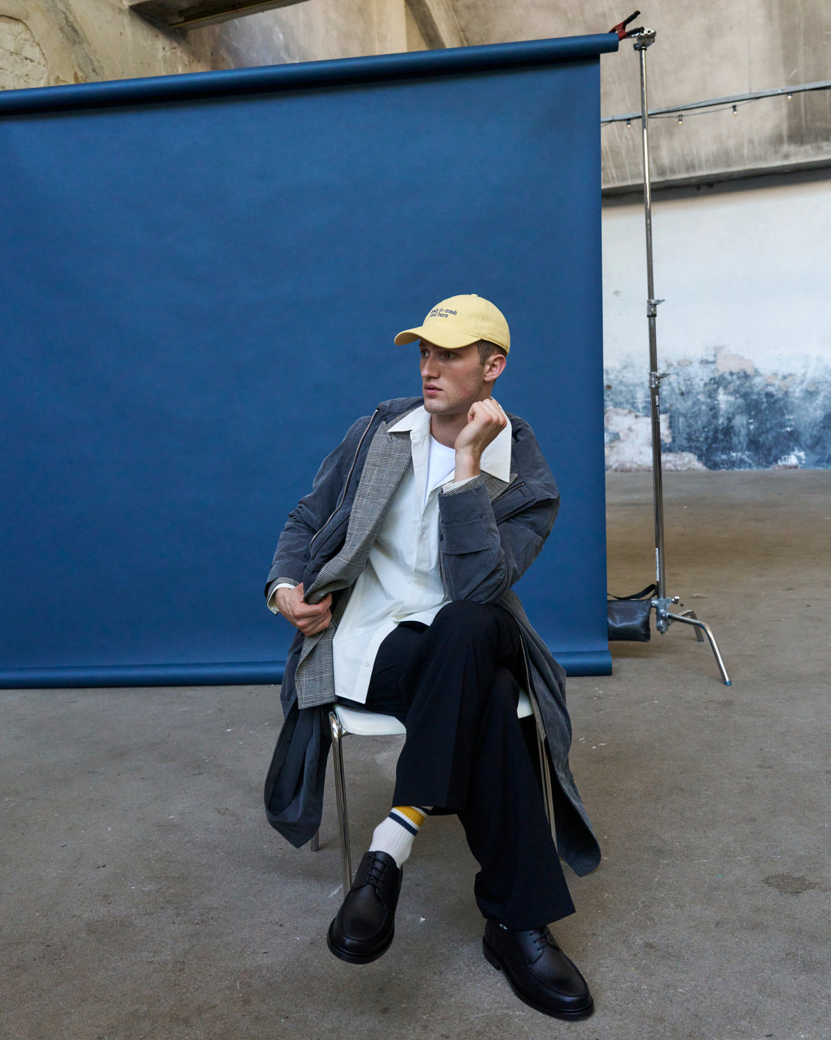 Person seated against a blue backdrop wearing the butter yellow Artless Ready To Crash And Burn organic cotton dad cap and a grey coat